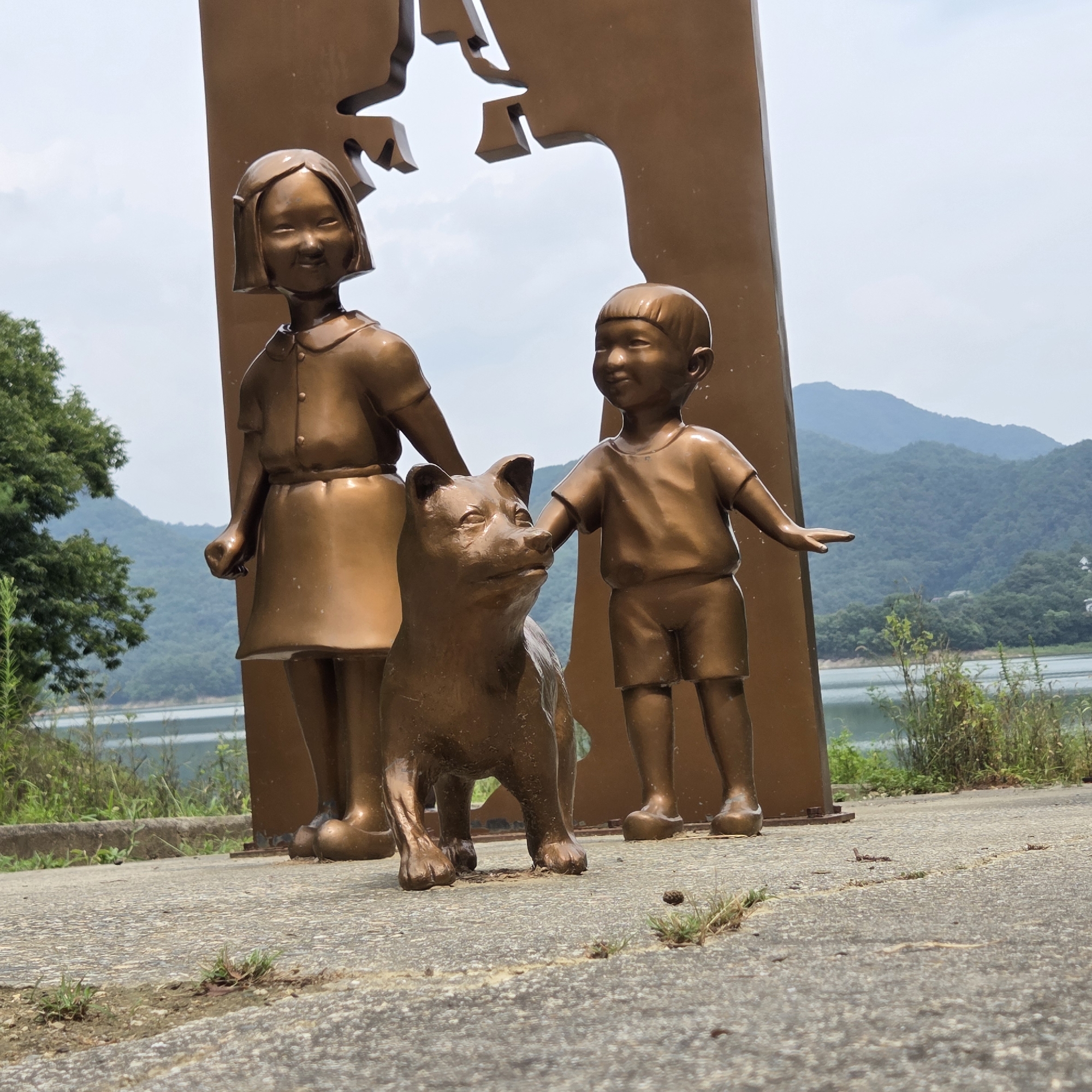This is a medium shot, eye-level, outdoor, full-body photograph of a bronze statue of two children and a dog. The statue is positioned in front of a large, abstract, bronze cutout of a silhouette that resembles human profiles. The girl on the left is wearing a dress and has short, bobbed hair. The boy on the right is wearing shorts and a t-shirt. Both children have their heads turned to the left, looking at the dog. The dog, also a bronze statue, is standing between the children, looking to its left. The statue is made of weathered bronze, with a slightly greenish patina in some areas.
The background features a body of water, possibly a lake or reservoir, with lush green hills and mountains rising behind it. The sky is overcast with some clouds. The ground in the foreground is a paved surface, likely concrete or asphalt, with patches of grass and dirt. The overall scene suggests a park or outdoor memorial site located in Gapcheon-myeon, South Korea, given the natural landscape. The lighting is diffused, indicating it is daytime.