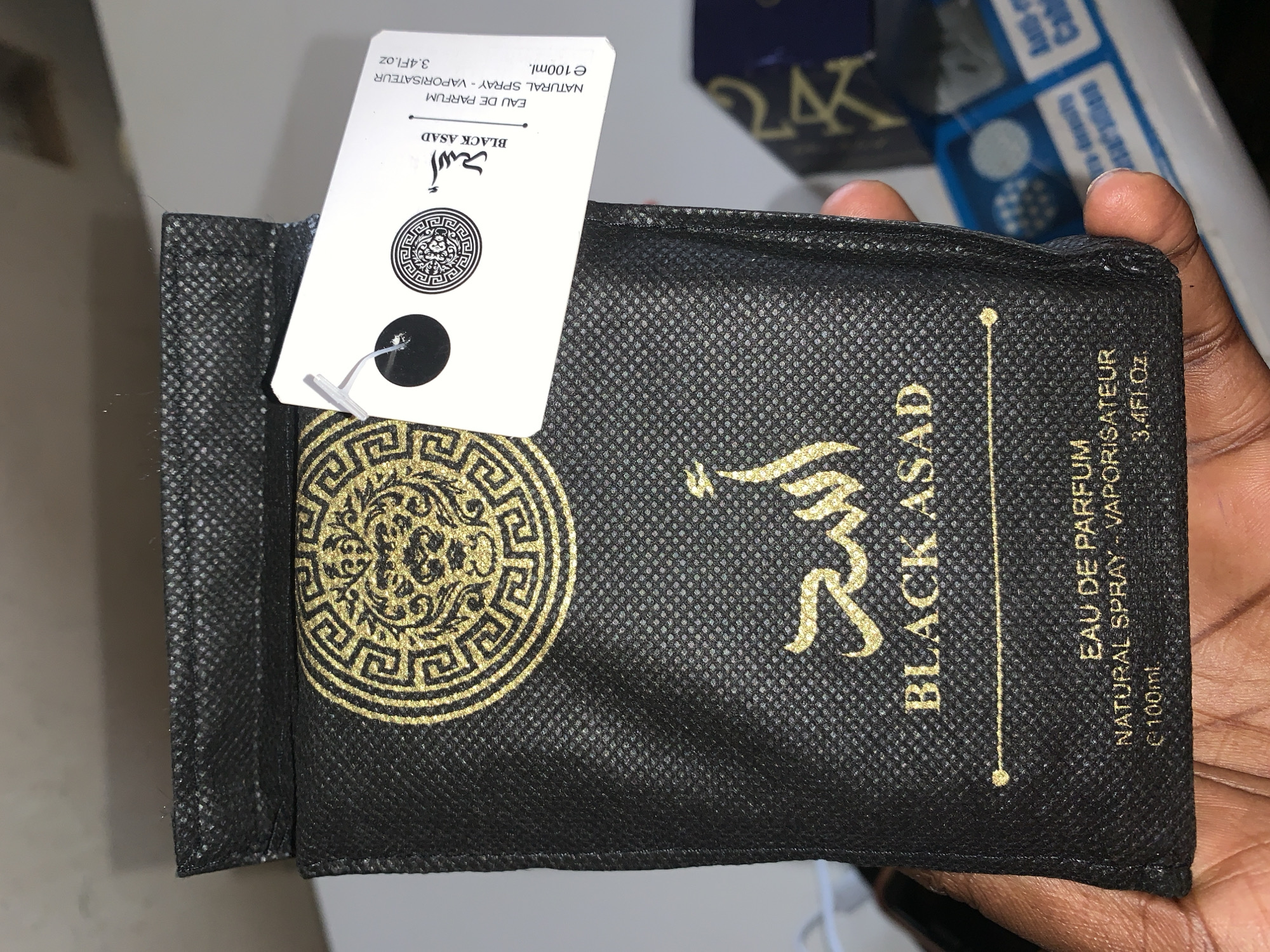 The image displays a close-up of a black, textured fabric pouch, likely containing a perfume bottle, held by a person's hand. The pouch features elaborate gold detailing, including the brand name "BLACK ASAD" in both English and elegant Arabic script ("أسد"). A circular gold emblem, resembling a classical Greek key or labyrinth pattern, adorns the lower left side of the pouch. Printed vertically along the right side of the pouch are the words "EAU DE PARFUM," "NATURAL SPRAY - VAPORISATEUR," "e 100ml.," and "3.4Fl.oz."
A white rectangular tag is attached to the top left of the pouch with a small clear plastic fastener. This tag replicates the same product information: "أسد," "BLACK ASAD," "EAU DE PARFUM," "NATURAL SPRAY - VAPORISATEUR," "e 100ml.," and "3.4Fl.oz."
A hand with a dark skin tone is visible on the right side of the frame, gently gripping the pouch. The background is softly blurred, revealing parts of other packaging or boxes, including a dark box with the gold text "24K" and elements of blue and white packaging, suggesting an indoor setting. The activity depicted is simply the holding or display of the product. There are no specific visual cues to determine the time of day, weather, or the precise location within New Stadium, Nigeria, beyond being an indoor environment. Emotions are not discernible.
Visible text includes: "أسد", "BLACK ASAD", "EAU DE PARFUM", "NATURAL SPRAY - VAPORISATEUR", "e 100ml.", "3.4Fl.oz" (repeated on both the pouch and the tag), and "24K" in the background.