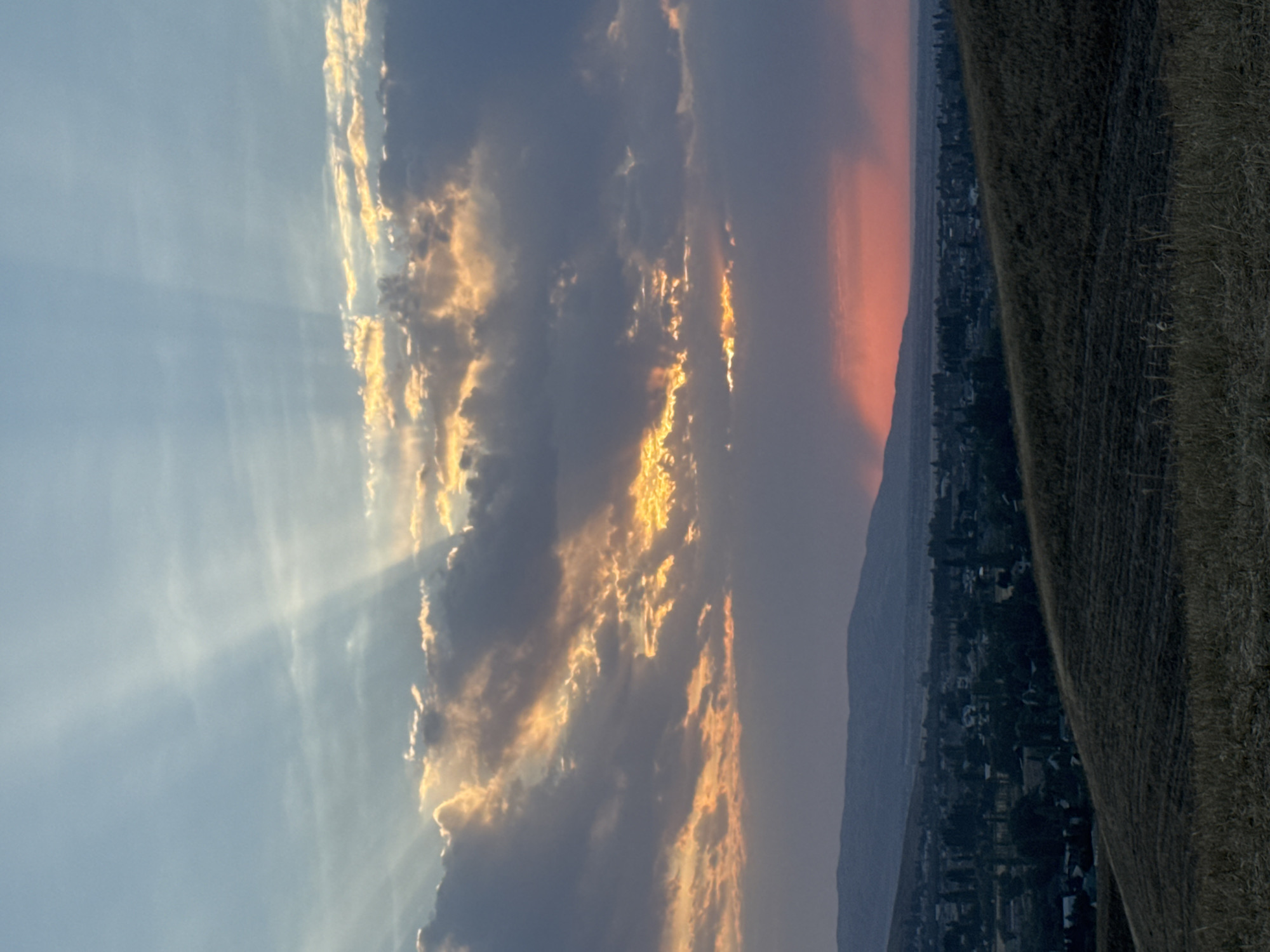 A high-angle, wide shot captures a twilight scene of a rural town nestled in a valley beneath rolling hills and mountains. The sky is a dramatic canvas of blues, grays, and oranges, with sunbeams piercing through scattered clouds. A vibrant orange glow on the horizon suggests the last moments of sunset. The foreground features a dry, grassy hillside, subtly textured by what appears to be cultivated fields. Beyond the hill, a town with clustered buildings and trees is visible, fading into the dusky landscape. A range of mountains forms a backdrop to the town, their peaks softened by the dimming light. The overall atmosphere is tranquil and expansive.