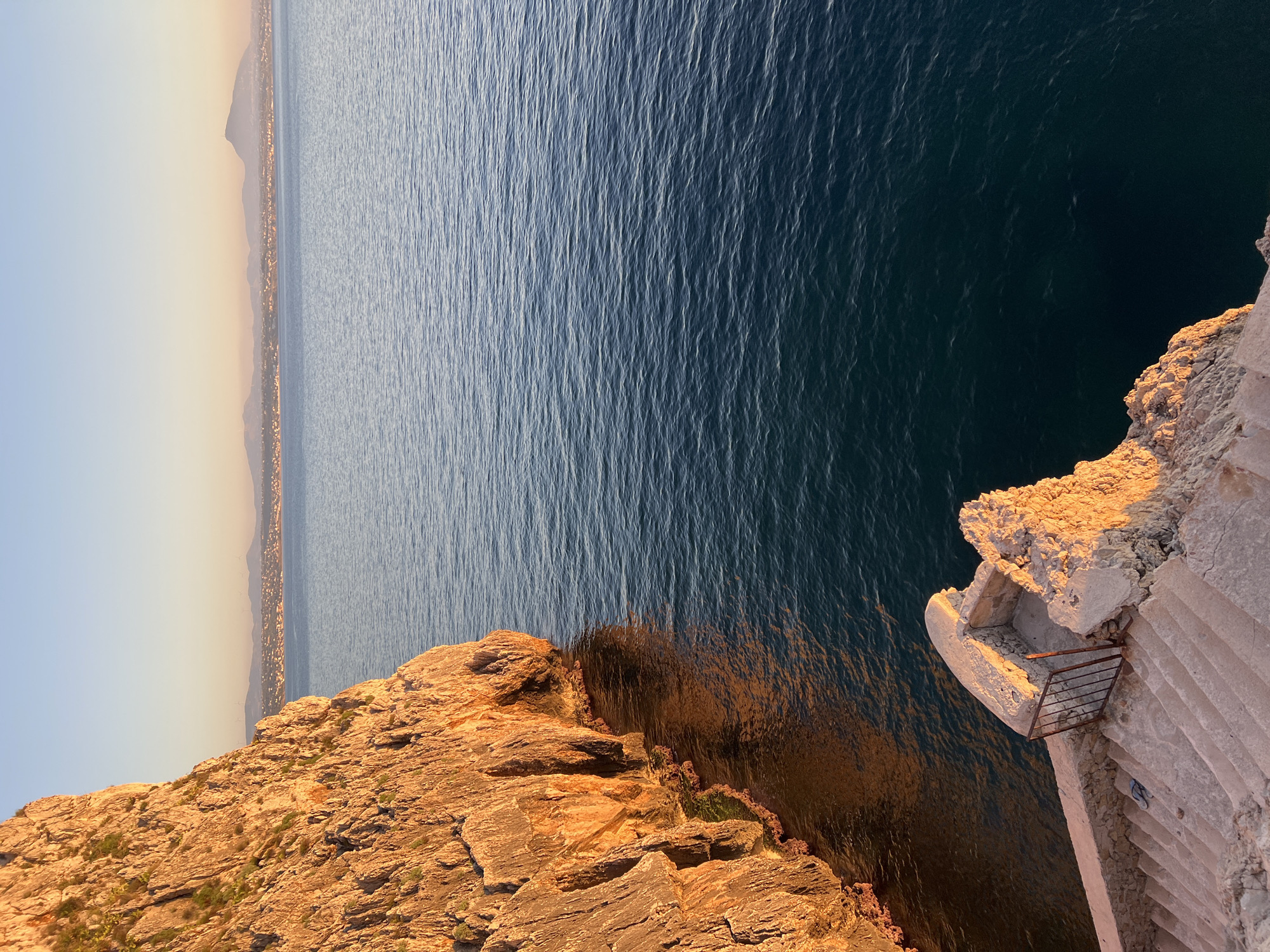 The image depicts a scenic view from a cliffside overlooking the sea at sunset. The sun is low on the horizon, casting a warm golden light on the rocky cliff face on the left side of the frame. The water is a deep blue, with gentle ripples on its surface. In the distance, a coastline is visible, dotted with buildings and illuminated by the setting sun. A stone staircase with a metal railing leads down from the foreground towards the sea. The overall scene conveys a sense of tranquility and natural beauty. The location is identified as Terrasini, Italy.