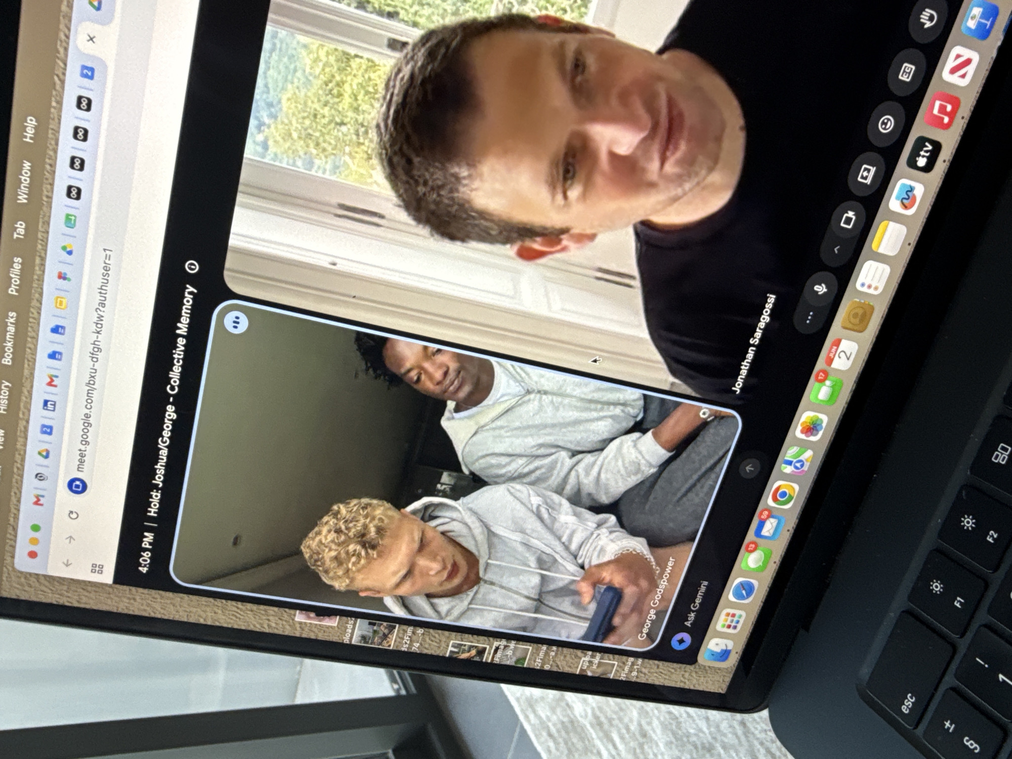 The image displays a close-up, angled view of a MacBook laptop screen, primarily showing a Google Meet video conference in progress. The reflection of the laptop user, a man with short, dark hair wearing a black t-shirt, is visible on the screen, looking directly towards the viewer. His video tile on the Google Meet call also shows his name as "Jonathan Saragossi".
The main video call window features a smaller tile with two participants, identified as "George Godspower". One young man with curly blonde hair wears a grey hooded sweatshirt and is looking down at a phone he holds. Behind him, another young man with dark hair wears a light-colored long-sleeved shirt.
The Google Meet call is titled "Hold: Joshua/George - Collective Memory" and indicates the time as "4:06 PM". An "Ask Gemini" button is also visible. The browser window above the video call has numerous tabs open, including "History", "Bookmarks", "Profiles", "Tab", "Window", "Help", Gmail, and LinkedIn, along with the Google Meet URL.
The laptop's dock is visible along the bottom of the screen, displaying various application icons such as Safari, Mail (with 59 unread messages), Messages (13 unread), Calendar (showing June 2), Google Chrome, Photos, and Apple TV. A partial view of the keyboard, including the "esc", "F1", and "F2" keys, is visible at the bottom right.
The setting appears to be indoors, with bright daylight coming through a window behind the laptop user, showing greenery outside. While the contextual location is Dubai, United Arab Emirates, there are no specific visual cues within the image to confirm this.