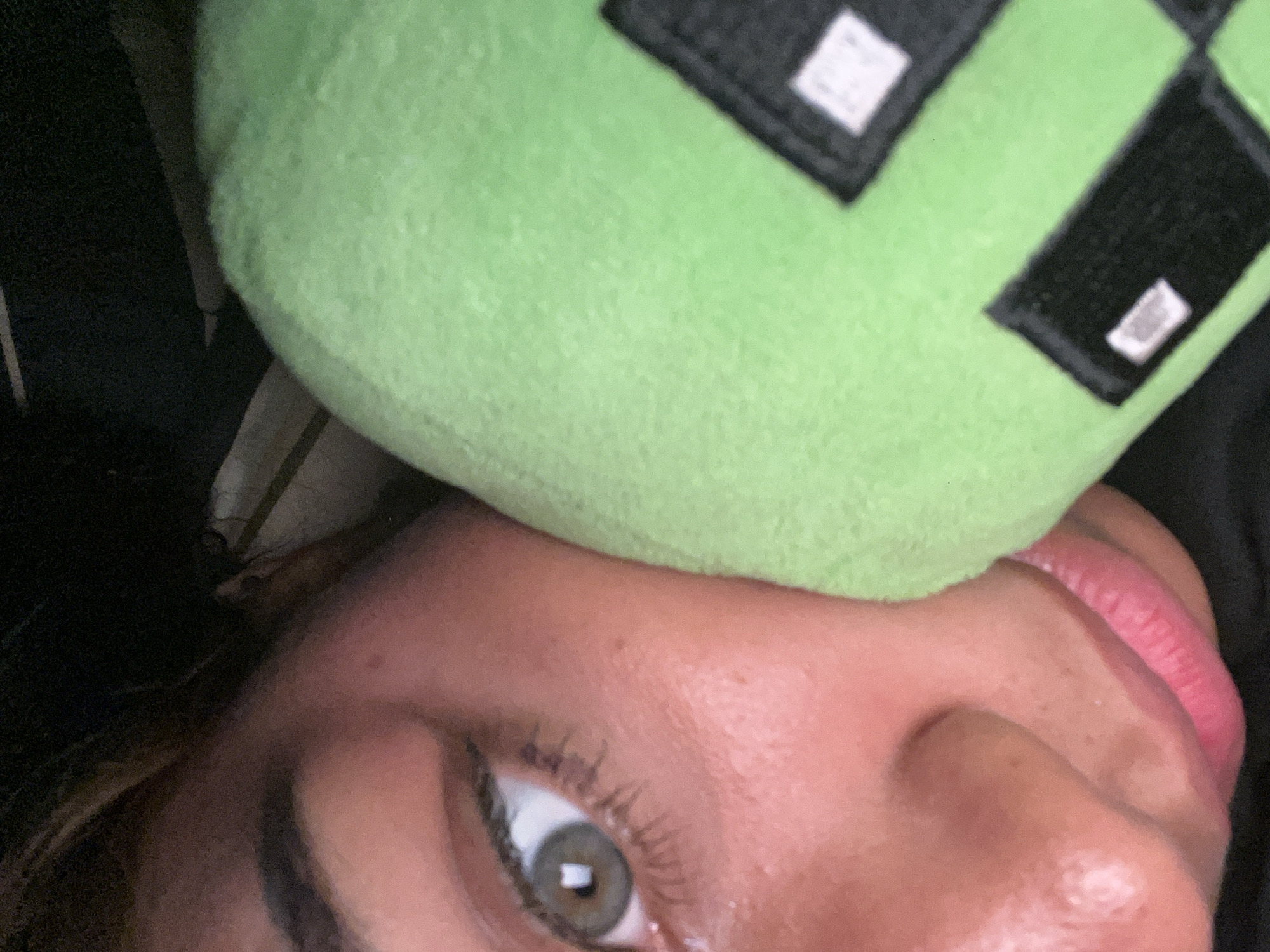 A close-up, tilted photograph captures a portion of a person's face with a bright green plush toy resting on their head. The person's left eye, which appears light-colored (possibly hazel or green) with long eyelashes, is open and directed towards the viewer. Part of their nose and pink-toned lips are also visible. The skin has a warm, smooth texture.
The prominent plush toy is a vibrant green and features black, pixelated square shapes with small white highlights, characteristic of a Minecraft Creeper. The toy has a soft, slightly fuzzy texture. A dark, out-of-focus background is visible to the left, suggesting an indoor setting with subdued lighting. The composition implies a relaxed or casual pose, possibly lying down. No discernible text is visible on the plush toy; the white elements are design details. While the context places the image in Pampatar, Venezuela, there are no specific visual cues within the photo to indicate this location.