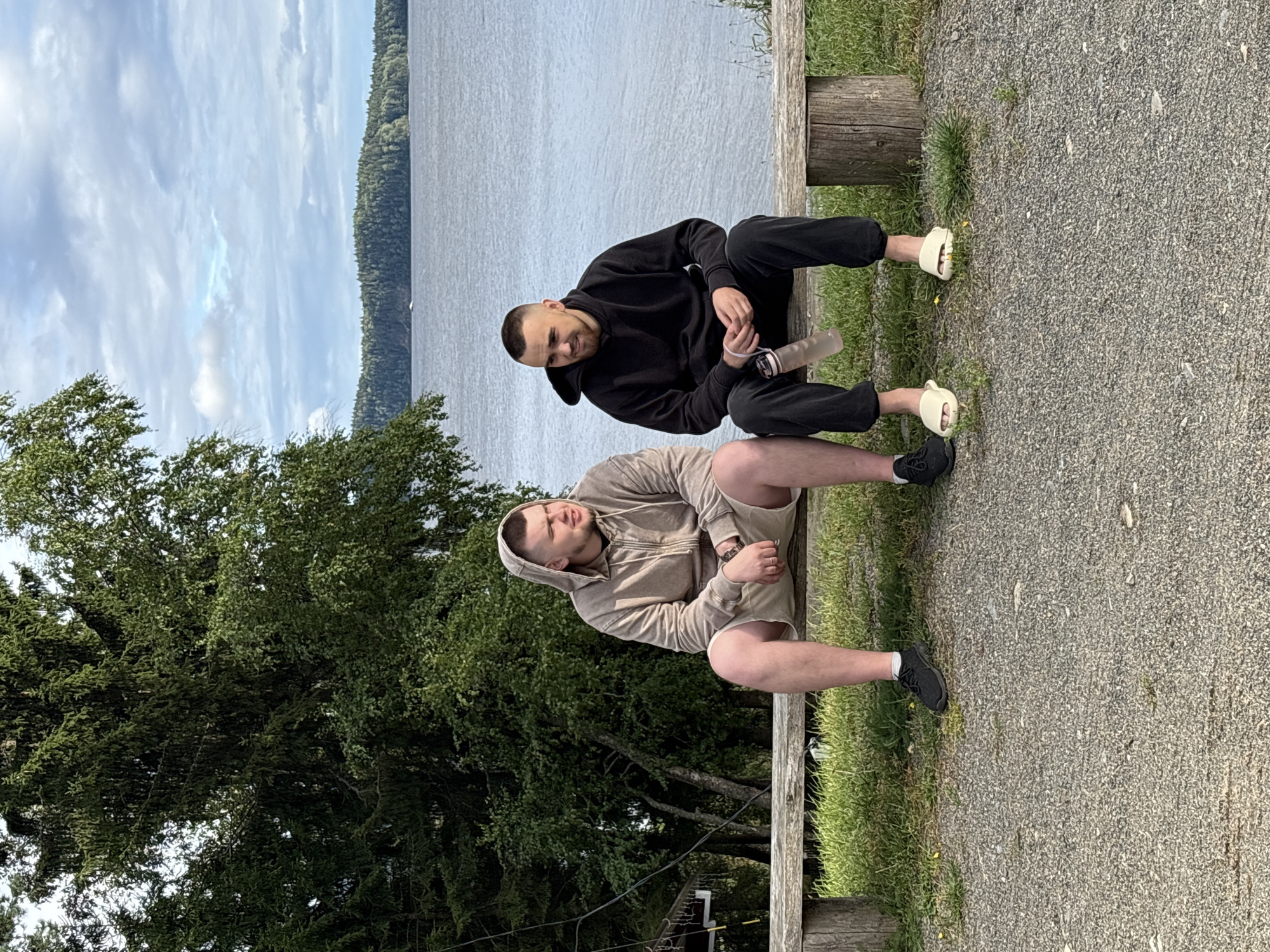 In Rautalakhti, Russia, two men are seated on a rustic wooden bench by a body of water. The man on the left wears a light brown hooded sweatshirt and light-colored shorts. The man on the right wears a black hooded sweatshirt and dark pants, with a water bottle with a pink strap resting beside him. They are positioned in front of a dense green forest that extends to the water's edge, under a partly cloudy sky. The ground in front of the bench is covered in gravel and sparse green grass.