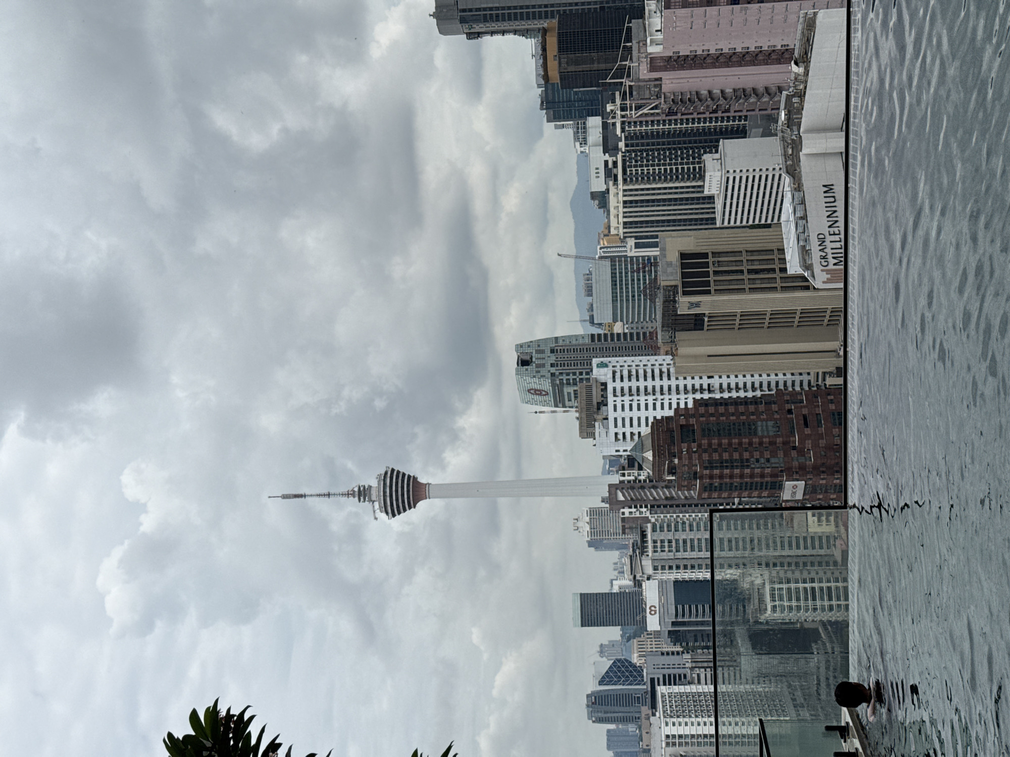 The image shows a cityscape with the Kuala Lumpur Tower (Menara Kuala Lumpur) prominently featured. The tower has a distinctive white and brown striped design and a tall, slender spire. In the foreground, water suggests a swimming pool or water feature, with a person visible in the water. The skyline is a mix of skyscrapers and buildings of varying heights and architectural styles, including the Grand Millennium Hotel. The sky is overcast with heavy cloud cover. The angle suggests the shot was taken from an elevated position, possibly a rooftop or high-floor balcony.