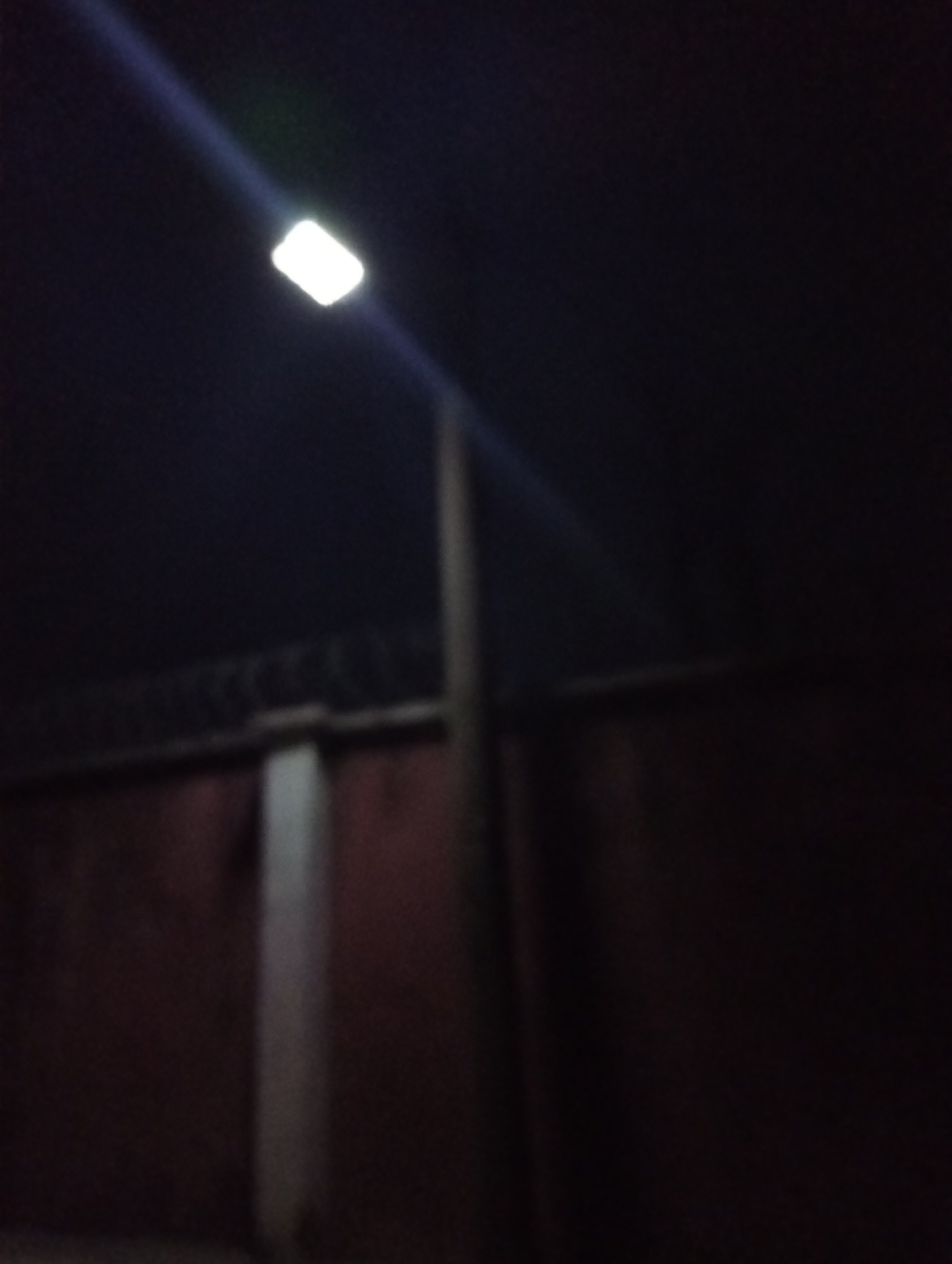 The image shows a dimly lit scene at night. A street light illuminates a section of a red brick wall topped with barbed wire. A concrete pillar stands adjacent to the wall. The location is potentially Jos, Nigeria, but this cannot be determined from the visual data alone. There are no visible people or actions. The scene is static and the main focus is on the light, wall, and barbed wire.