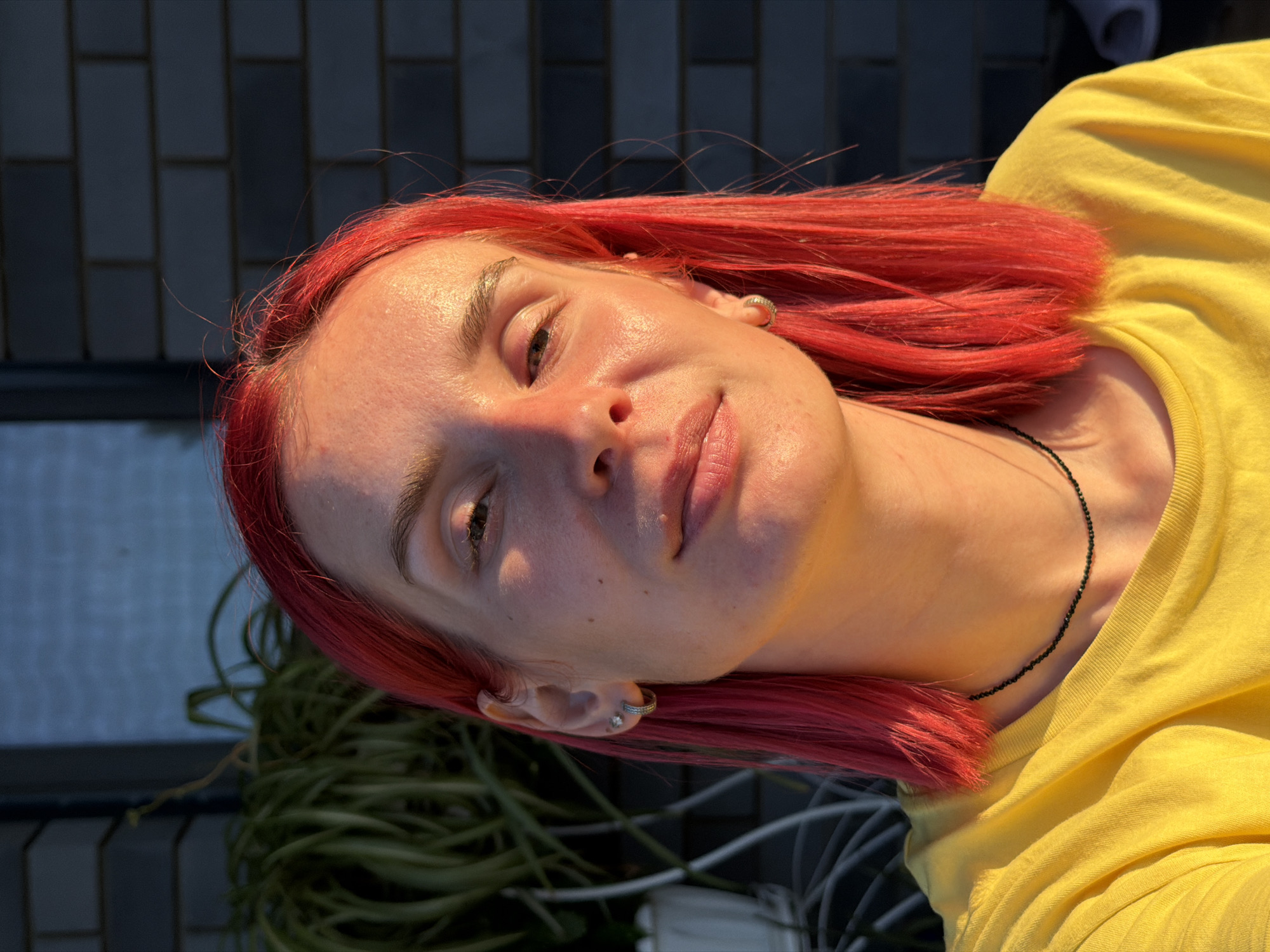This is a close-up, head-and-shoulders portrait of a young person with bright red hair, looking upwards. The subject is wearing a bright yellow t-shirt and a thin black beaded necklace. Sunlight casts a shadow across the left side of their face, highlighting the contours of their nose and cheekbone, and the left side of their forehead. Their eyes are open, and their lips are slightly parted. The person has a small stud earring in their left ear and a hoop earring with a small gem in their right ear.
In the background, dark, rectangular tiles form a wall or surface. To the left of the subject, a tangled mass of green, vine-like objects is visible, possibly plants or wires. The lighting suggests it is daytime, with strong sunlight.
The image appears to be taken outdoors. The subject's pose and the direct sunlight imply a casual, perhaps relaxed or contemplative moment. The overall mood is bright and warm, due to the sunlight and the yellow shirt. There is no visible text in the image.