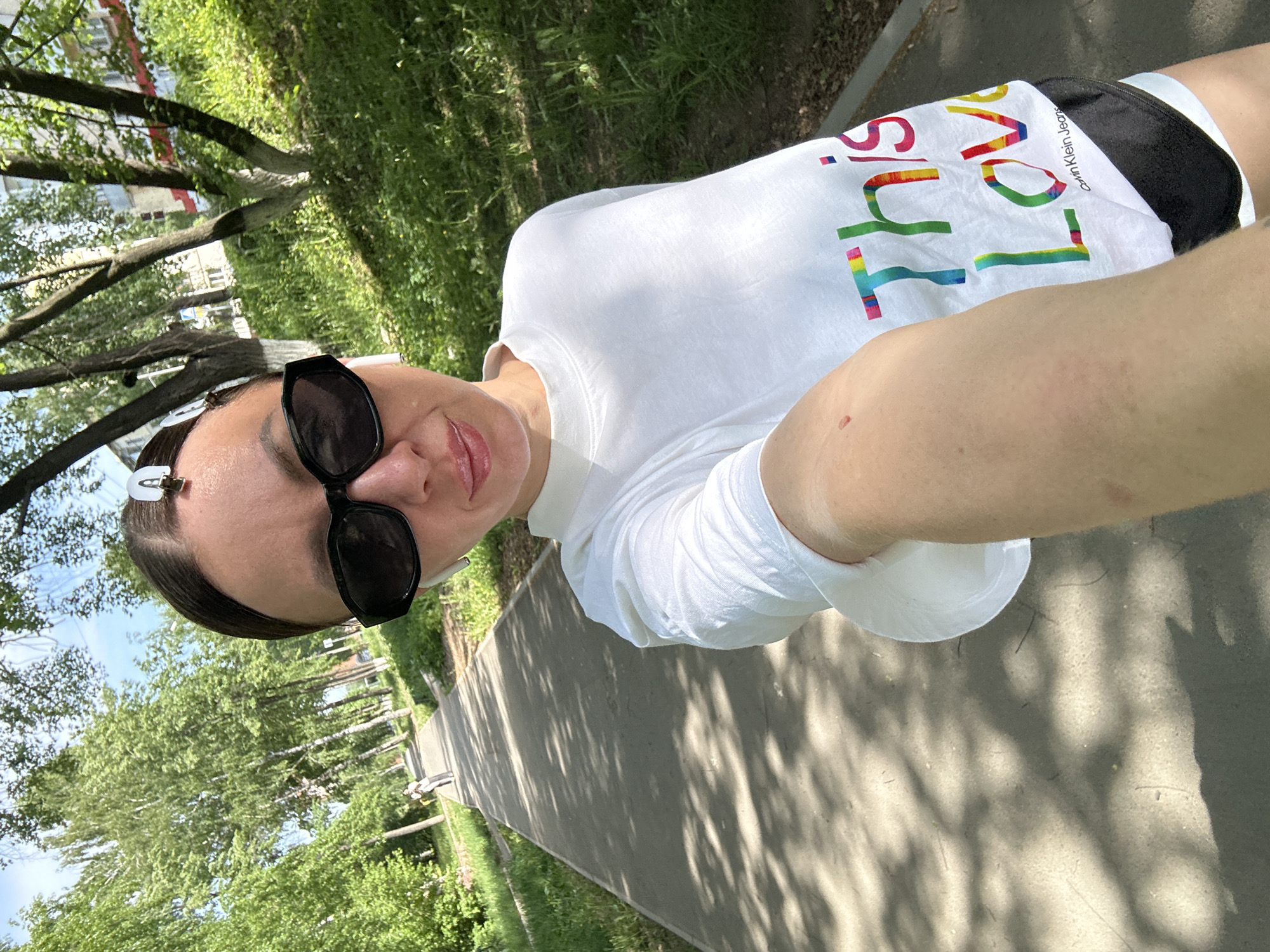 A selfie captures a woman wearing sunglasses, a white t-shirt with "THIS LOVE" printed in rainbow colors, and black shorts. Her hair is pulled back with a clip. The photo is taken outdoors, likely in Votkinsk, Russia, given the context. The background includes trees, grass, and a paved walkway. Sunlight filters through the trees, casting shadows on the ground. The image suggests she is either walking or standing still on the path.