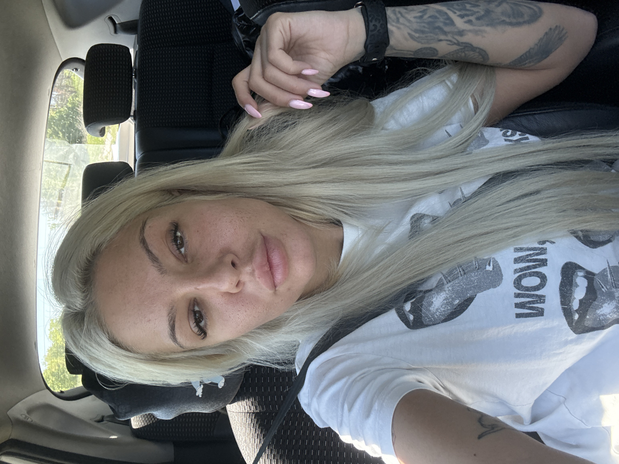 A woman is in the driver's seat of a car. She is wearing a white t-shirt and has long blonde hair. The car is parked on a street in Pesnopoy, Bulgaria. The woman is looking at the camera.