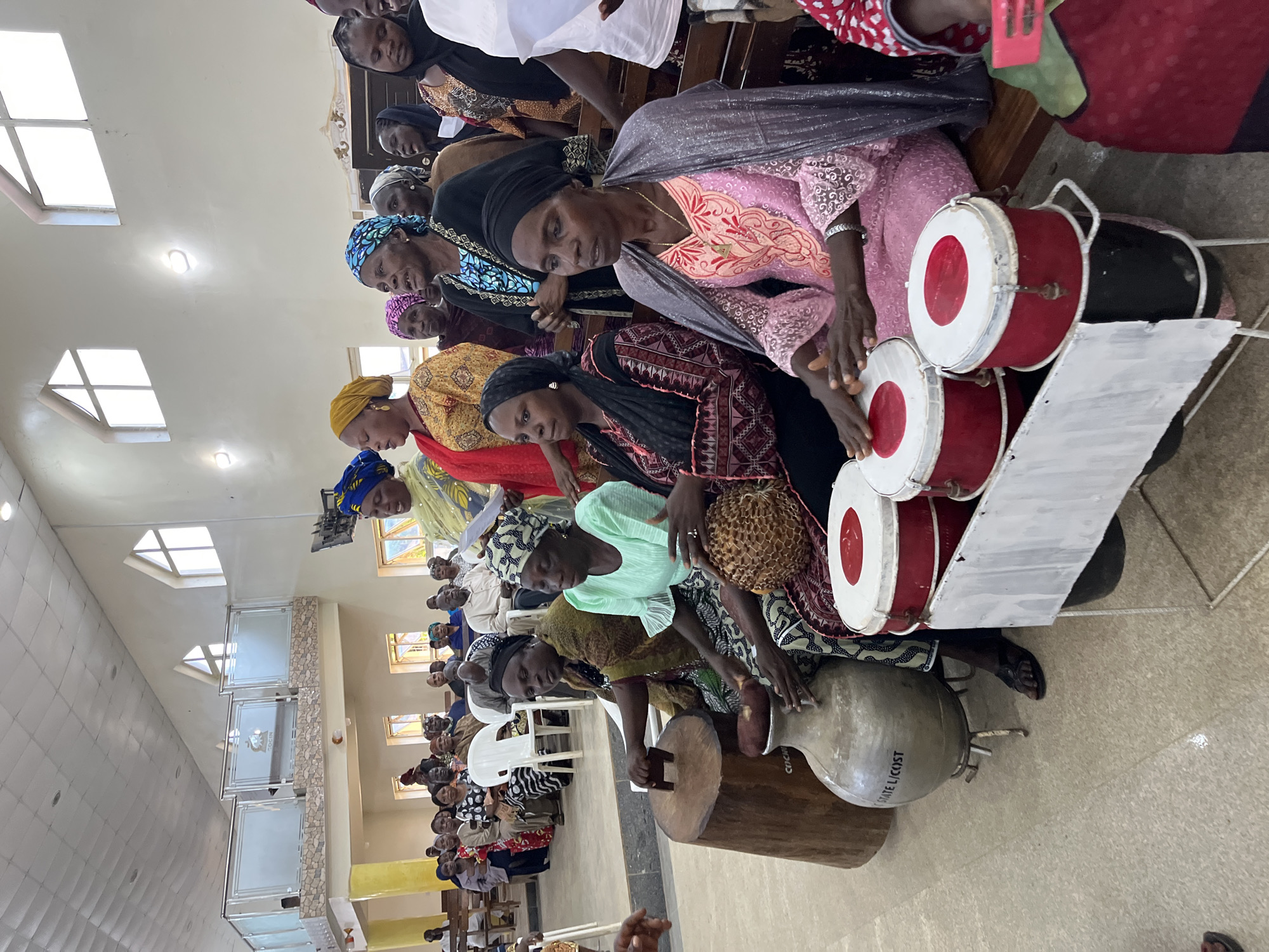 A low-angle shot shows a gathering of people inside a building. The scene is centered on a group of women playing drums, possibly a religious or cultural event. The location is Jos, Nigeria, as indicated by text on one of the instruments.
There are several women seated in rows, and they are arranged in the foreground. The women are dressed in colorful traditional Nigerian clothing, some with headscarves or wraps. They are actively engaged in playing a variety of drums, some with red and white accents.
Behind the women, rows of people are seated. They appear to be an audience or participants in the event.
The building has architectural features of a modern church, including windows and light fixtures. The room is illuminated by natural light and overhead lighting.