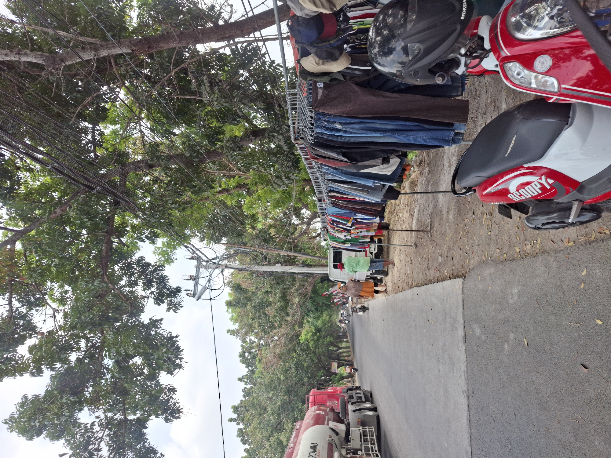 This image depicts a roadside scene in Sidoharjo, Indonesia, likely a commercial area with street vendors. On the left, a red Pertamina tanker truck is parked on the asphalt road. To the right of the road, a clothing rack displays a variety of garments including shirts, pants, and jackets. Several hats are also visible hanging on the rack. In the foreground, two red and white scooters are parked. A person in a green shirt and a person in an orange hijab are standing near a white pick-up truck, possibly a vendor and a customer. The background features a line of trees and a power pole with numerous wires. The ground is a mix of dirt and scattered leaves.
