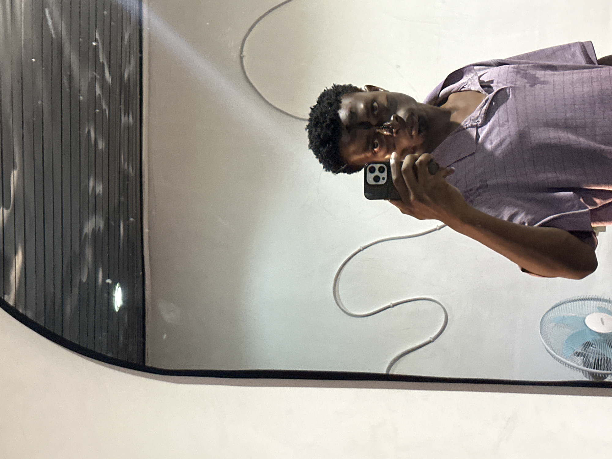 A person is taking a selfie in a mirror. The person is wearing a purple patterned shirt and holding a smartphone. The mirror reflects a white wall with a white cable running along it, and a white fan with blue blades. The location is possibly Bukuru, Nigeria.