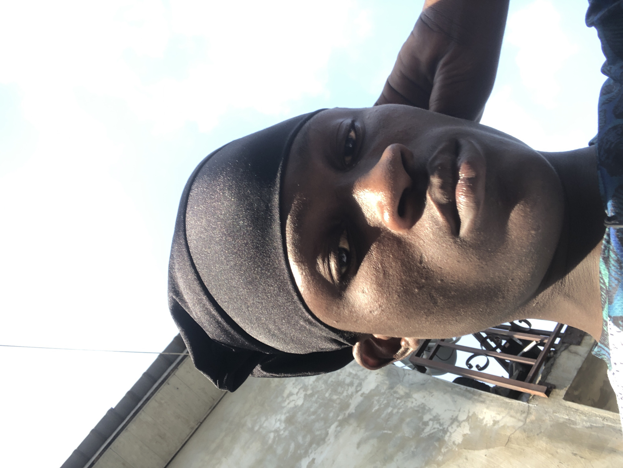 A close-up, low-angle shot captures a young Black man from the shoulders up, looking directly at the camera. He is wearing a black do-rag and a blue and green patterned t-shirt. His right hand is raised to the back of his head, and his left ear is visible. Sunlight casts shadows across his face, highlighting his features and the texture of his skin.
Behind him, a concrete wall with rough texture and visible cracks is on the left. A section of a building with a tiled roof is visible in the upper left corner. A metal railing with decorative scrollwork separates the wall from the sky. The sky is bright blue with some white clouds, suggesting a clear day.
The scene appears to be outdoors, possibly on a balcony or rooftop, given the vantage point and the presence of the railing and building structures. The overall impression is one of a casual, everyday moment captured under bright daylight. The location is identified as Ado, Nigeria, though no specific landmarks are identifiable within the image to confirm this beyond the provided context.