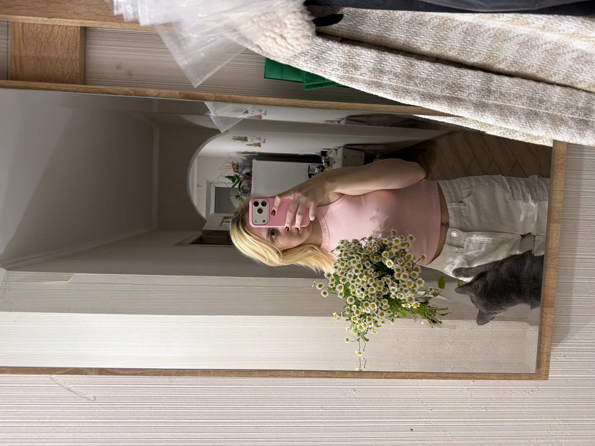 A young woman with blonde hair and a pink top is taking a mirror selfie in what appears to be a hallway or entryway in a residential setting in Cheltuitori, Moldova. She is holding a bouquet of white daisies and a pink smartphone. A gray cat is visible in the reflection, looking towards the bouquet. The environment includes a large mirror framed in wood, textured wallpaper, a wooden door frame with an arched opening in the background revealing a domestic interior, and clothing hanging on the right side of the frame. The lighting suggests it is indoors, possibly in the evening.