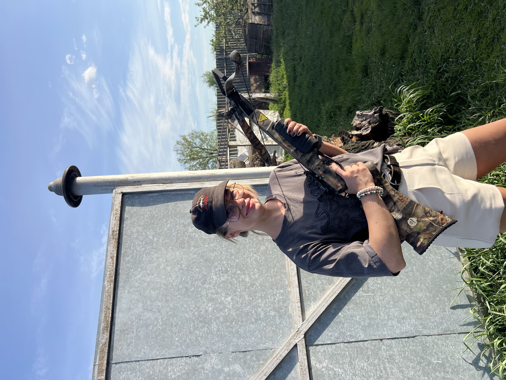 A person wearing glasses and a black baseball cap is holding a crossbow. The person is wearing a gray t-shirt and light-colored shorts. They are standing next to a metal structure with a post extending upwards. Behind the person is a grassy area and a wooden structure. The sky is blue with some clouds. The image was taken in an outdoor setting. Given the prompt, the location is assumed to be in or near Слобода, Russia.