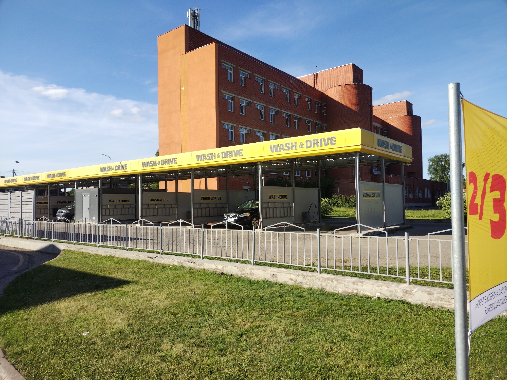 The image depicts a car wash facility with multiple bays, identified by "WASH & DRIVE" signage, located in Daugavpils, Latvia. A modern, brick building with multiple windows stands in the background, with visible cell towers on its roof. In the foreground, a bright yellow banner with the text "2/3" and some unreadable Latvian text is partially visible, attached to a metal pole. A dark-colored car is present in one of the car wash bays. The area is bordered by a metal fence and a strip of grass. The sky is clear and blue with a few scattered clouds.