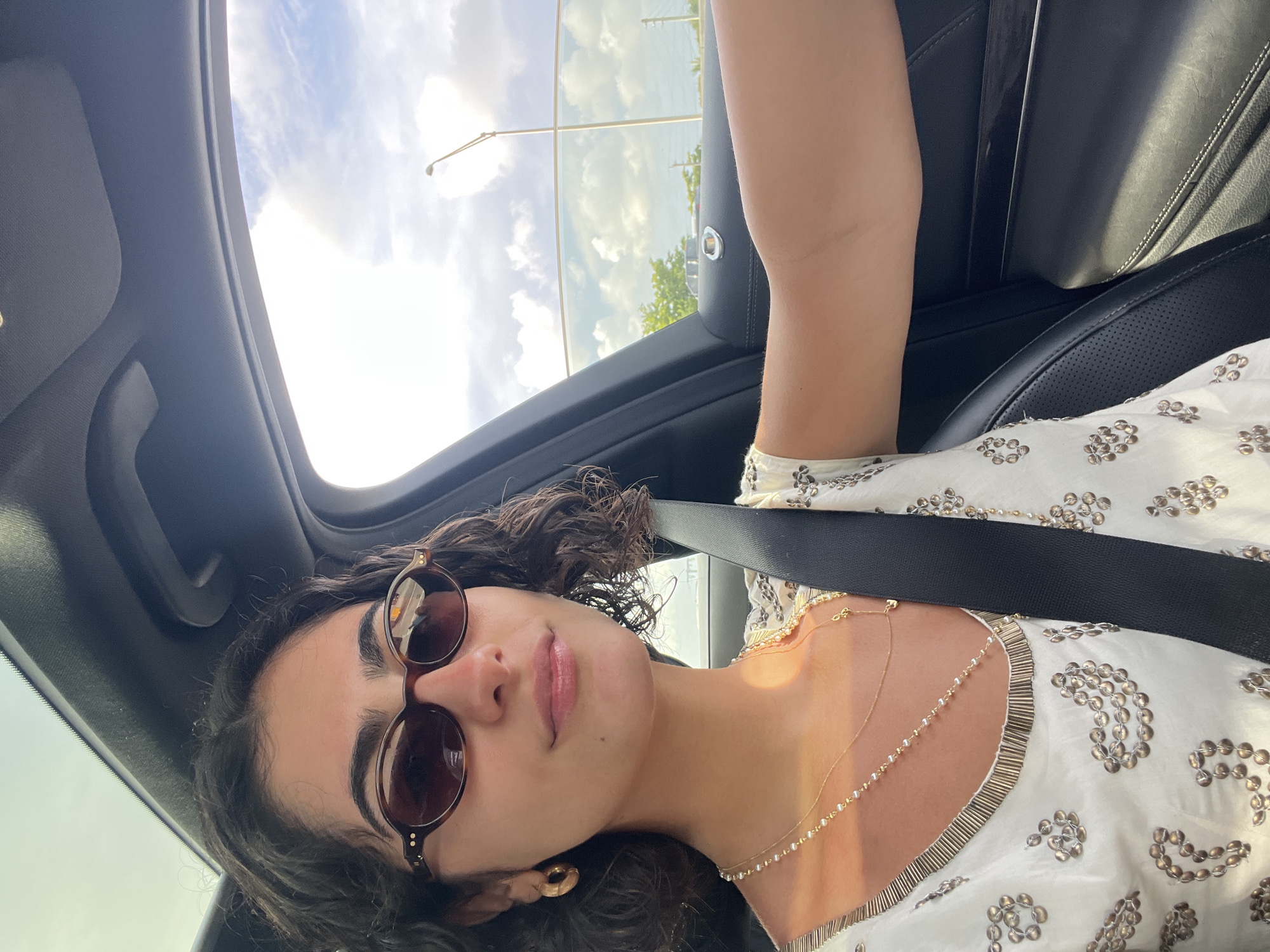 A woman is inside a car, likely in Miami, given the sky and palm trees visible through the sunroof. She is wearing sunglasses, a light-colored top with embellishments, and a gold necklace. A seatbelt is visible across her chest. The interior of the car is dark, with leather seats. The image is taken from a tilted perspective.