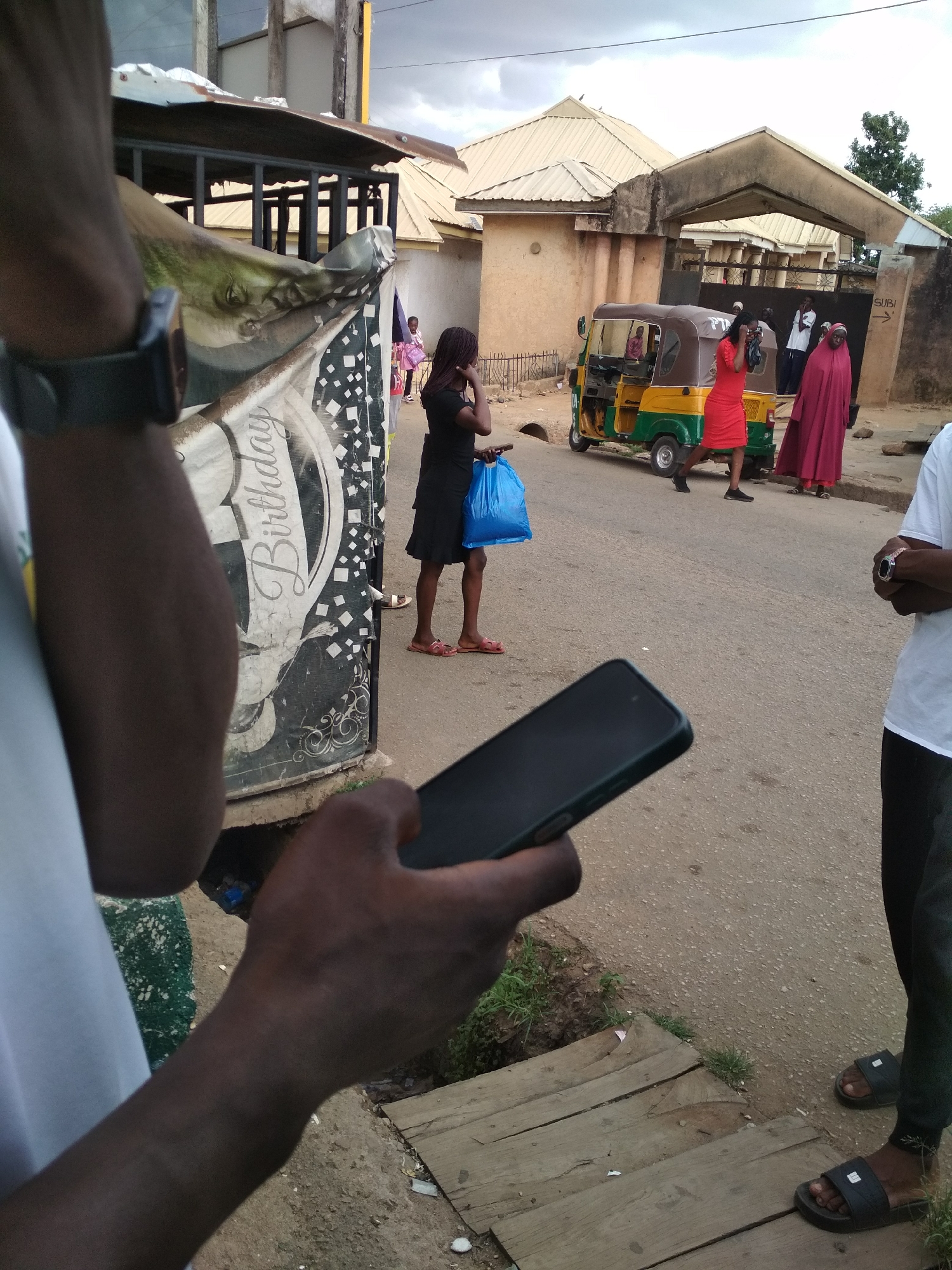 The image shows a street scene in Gura Top, Nigeria. In the foreground, a person holds a smartphone. A woman in a black dress stands on the street, holding a blue plastic bag. Further down the street, a yellow and green auto-rickshaw is parked near a building entrance. Two women, one in a red dress and the other wearing a maroon hijab, stand near the entrance. Another person wearing black pants and a white shirt is standing on the right-hand side of the image. The street is paved, with wooden planks covering a drainage channel in the foreground. An advertisement containing the word "Birthday" is visible on the left side of the image. The sky appears overcast.