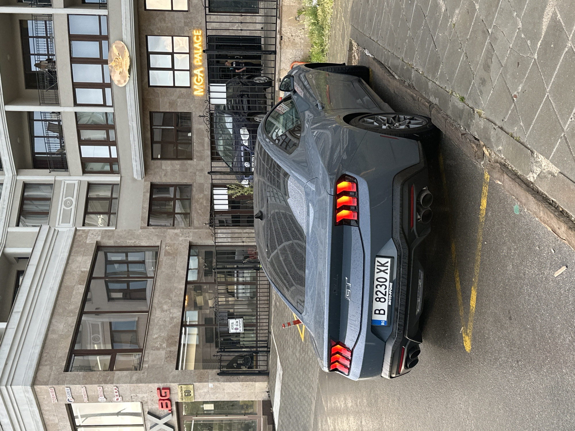 The image shows a gray Ford Mustang parked on a street next to a building. The car has Bulgarian license plates reading "B 8230 XK." The building appears to be commercial, with the words "MGA PALACE" visible, and also has signs for Toshiba, Gree, General, Bosch, and XEG brands. The building has a tan exterior with brown window frames. The scene suggests a location in Varna, Bulgaria due to the Bulgarian license plate and the presence of Cyrillic text on a sign ("Изход"). The car is parked alongside the curb with yellow painted parking lines. The image appears to be taken during the day.