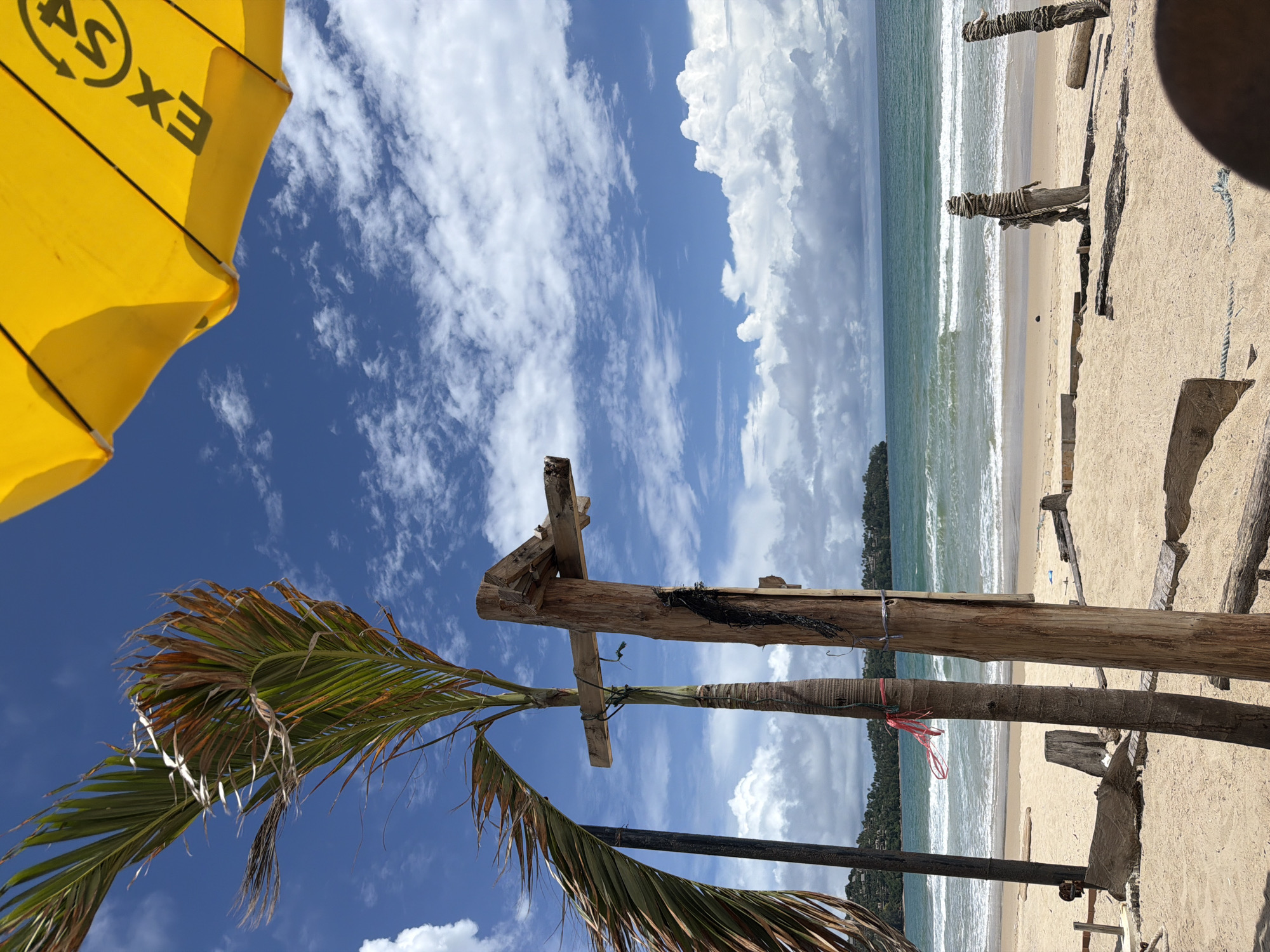 The image depicts a tropical beach scene in Choeng Thale, Thailand. In the foreground, weathered wooden posts and beams are arranged on the sand, suggesting the remnants of a structure or some form of beachside construction. A palm tree with fronds that appear dried and windblown stands to the left of the wooden elements. A yellow umbrella, partially visible at the top right of the frame, has some text and a logo on it, possibly indicating a brand or service.
The middle ground showcases a sandy beach that slopes gently towards the ocean. Waves are breaking on the shore, indicating a moderate sea state. The water is a turquoise blue, transitioning to a deeper blue as it extends towards the horizon. In the distance, a coastline with lush green hills and scattered buildings is visible. The sky is a vibrant blue, dotted with fluffy white cumulus clouds, characteristic of a tropical climate. The overall impression is one of a relaxed, perhaps slightly weathered, beach environment under a clear, sunny sky.