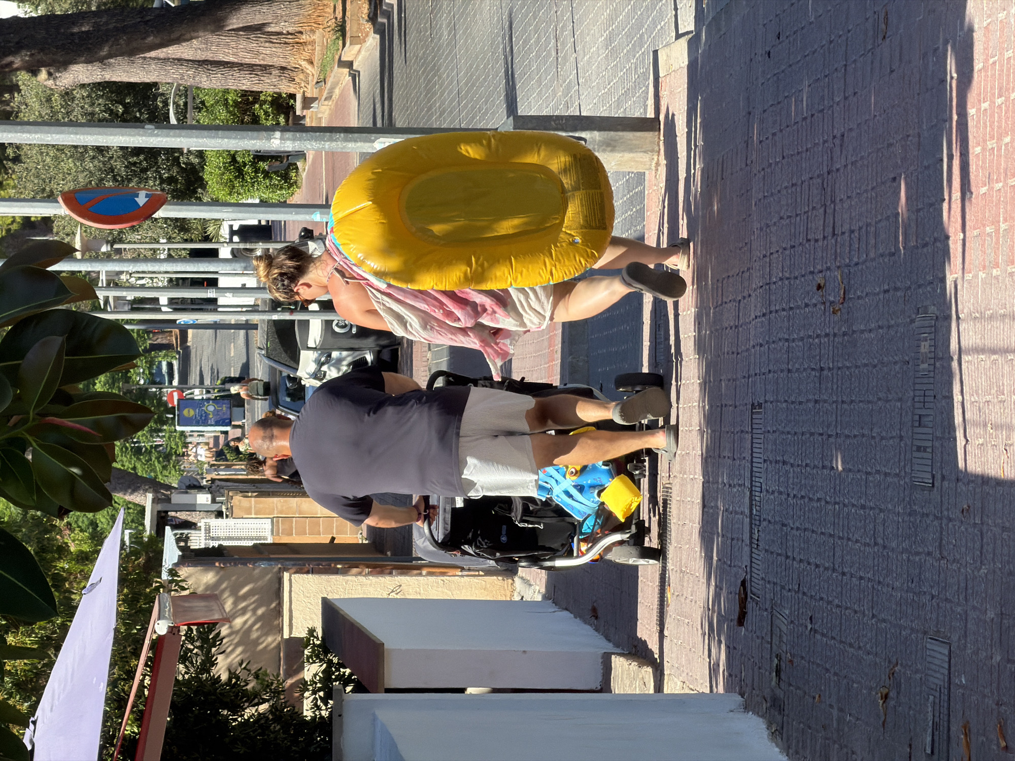 In Palmanova, Spain, a man wearing a dark t-shirt and white shorts pushes a stroller on a tiled sidewalk. A woman, her back to the camera, wearing a light-colored dress, carries a large yellow inflatable object on her shoulder and a towel draped over it. The sidewalk is lined with trees and buildings. A blue and red traffic sign with a white arrow is visible on a pole. The scene suggests a warm day, possibly for a beach outing.