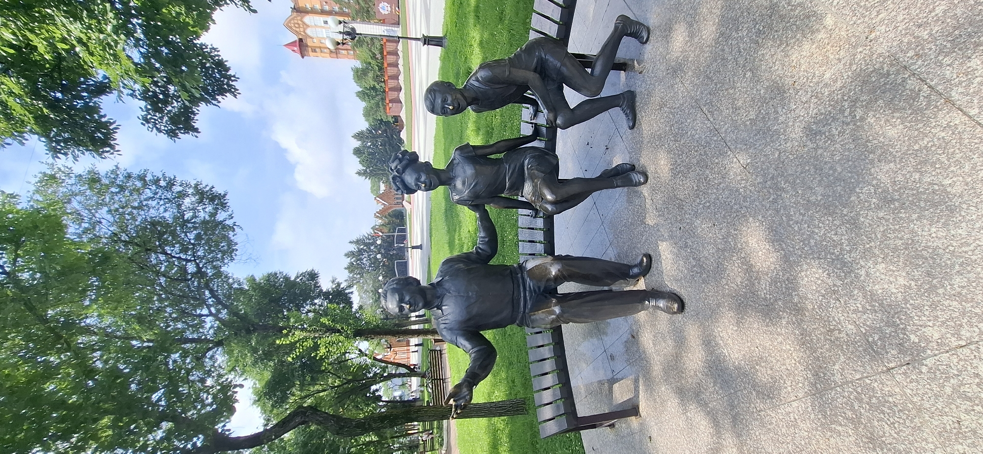 A bronze sculpture depicts three figures seated on a dark wooden bench in a park. On the left, an adult male in a collared shirt and trousers gestures outwards with his right hand. To his right sits a young girl with her hair styled in a bun, wearing a sleeveless dress. On the far right, a young boy kneels on the bench, looking towards the other figures. The sculpture is placed on a paved area, with shadows from overhead trees dappling the ground. In the background, a manicured lawn and trees are visible, leading to a street with a building featuring a red-roofed tower and lampposts. This scene is located in the city of Blagoveshchensk, Russia.