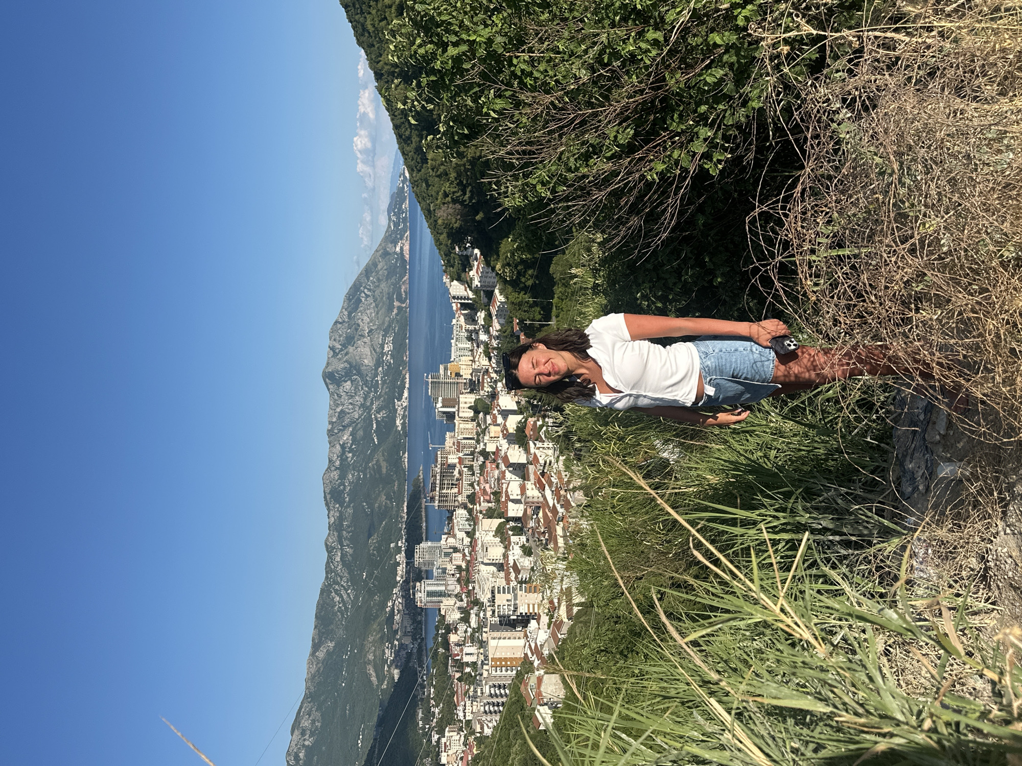 A woman stands in the foreground on a dirt path, surrounded by dry, thorny bushes and tall green grass, looking towards the camera with her eyes closed and a slight smile. She is wearing a white short-sleeved shirt and denim shorts. She holds a dark-colored phone in her right hand. Behind her, a panoramic view of the city of Budva, Montenegro, unfolds. Buildings with terracotta roofs and modern high-rises are clustered together, sloping down towards a blue bay. Mountains form a dramatic backdrop to the city, with a clear blue sky above. The sun is casting a warm light on the scene.