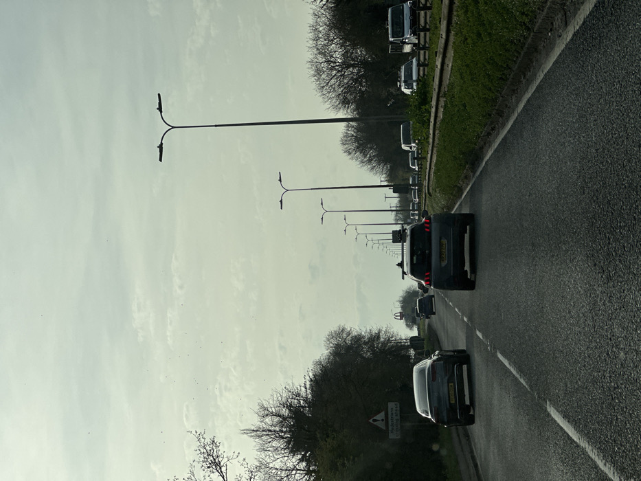 The image depicts an asphalt road scene in Basildon, United Kingdom, specifically on Laindon Link (B1007) heading northbound towards the Fortune of War roundabout and the A127 (Southend Arterial Road). The sky is overcast with visible cloud formations.
In the foreground, a dark grey Peugeot 3008 SUV, bearing the registration SR70 ZPE, is visible with roof racks, facing away from the viewer. Immediately behind it, a dark grey sedan with registration BG70 XOW is also traveling in the same direction. Multiple additional vehicles, including cars and vans, are present in the mid-ground and background, indicating moderate traffic flow.
On the left side of the road, a triangular warning sign with a red border and a black pedestrian symbol is visible, below which a rectangular white sign reads "Pedestrians Crossing Ahead". The left roadside features dense vegetation, primarily deciduous trees with bare branches or nascent foliage. A prominent yellow McDonald's 'M' arch logo is visible in the mid-distance, positioned to the right of the road's center line.
The right side of the road features a grass verge, a low metal barrier, and further bare-branched trees and sparse vegetation. A series of modern streetlights, each with a dual fixture, extends along both sides of the road into the distant background, indicating a major arterial route. No pedestrians are visible in the scene.