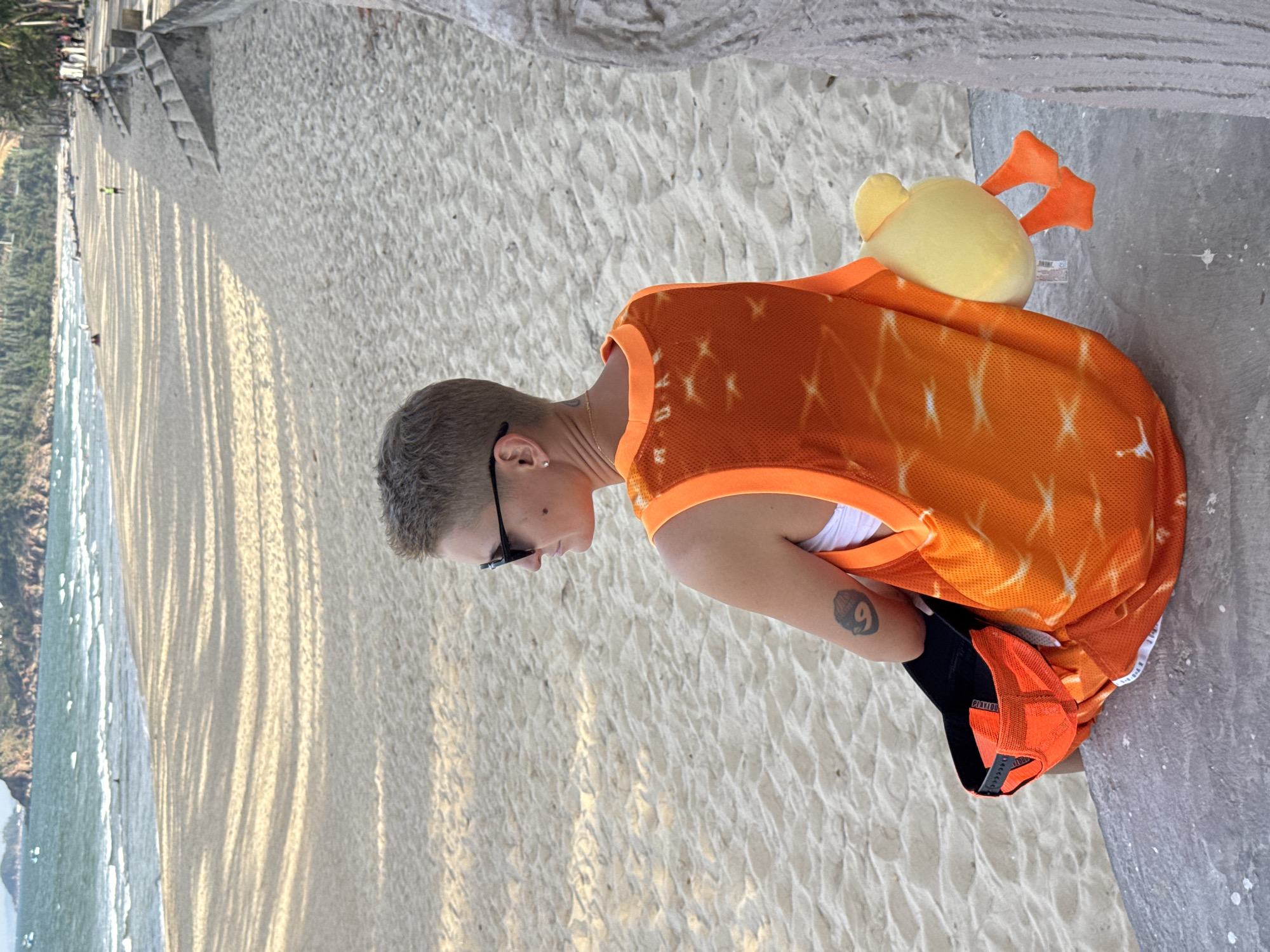 A person with short, blonde hair and sunglasses is sitting on a concrete ledge facing away from the camera, looking out at a sandy beach and the ocean. The person is wearing an orange sleeveless jersey over a white top, and a black baseball cap is tucked into their side. A yellow plush duck toy is attached to the back of the jersey. The beach is wide with light-colored sand and dappled shadows from unseen trees or structures. The ocean is a vibrant blue-green with small waves, and in the distance, hills covered in green vegetation rise from the coastline. The scene appears to be in a public area, possibly a promenade or park, along the beach in South Quy Nhon Ward, Vietnam, given the coastal environment and architecture.