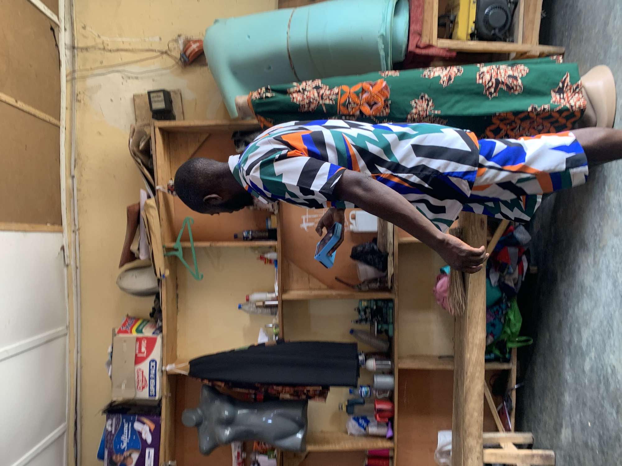 The image depicts an indoor scene, possibly a workshop or shop, in Jos, Nigeria. A man wearing a colorful, geometric-patterned shirt and shorts stands in the foreground, holding a small blue object in his right hand. He is facing away from the camera, towards a shelf unit. The shelves are wooden and appear to be stocked with various items, including bottles, containers, fabric, and a sewing machine. To the left of the man, a dark grey mannequin torso is visible. To his right, a mannequin is dressed in a green, floral-patterned dress. Behind the mannequins, a large, light blue cylindrical object is present. The walls of the room are painted a light yellow, and the ceiling is white with visible beams. The floor is a dark grey concrete. The overall impression is that of a busy, functional space, likely involved in textile work or retail.