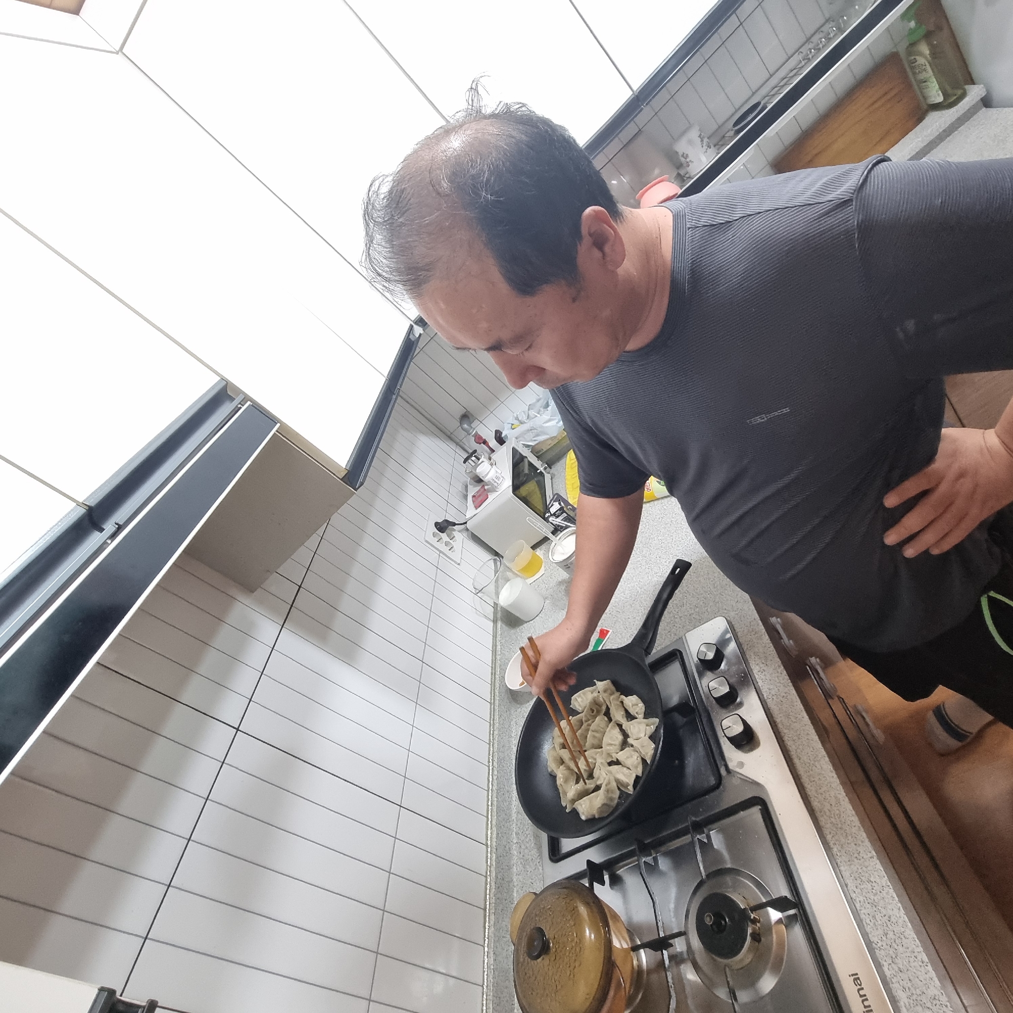 In a kitchen, an adult male with thinning gray hair is cooking dumplings in a frying pan on a gas stovetop. He is wearing a dark gray t-shirt. His right hand is holding chopsticks and is in the process of turning the dumplings in the pan. To the left of the pan is a brown pot with a lid. The kitchen has white tiled walls and a gray speckled countertop. The overall scene depicts a domestic cooking activity.