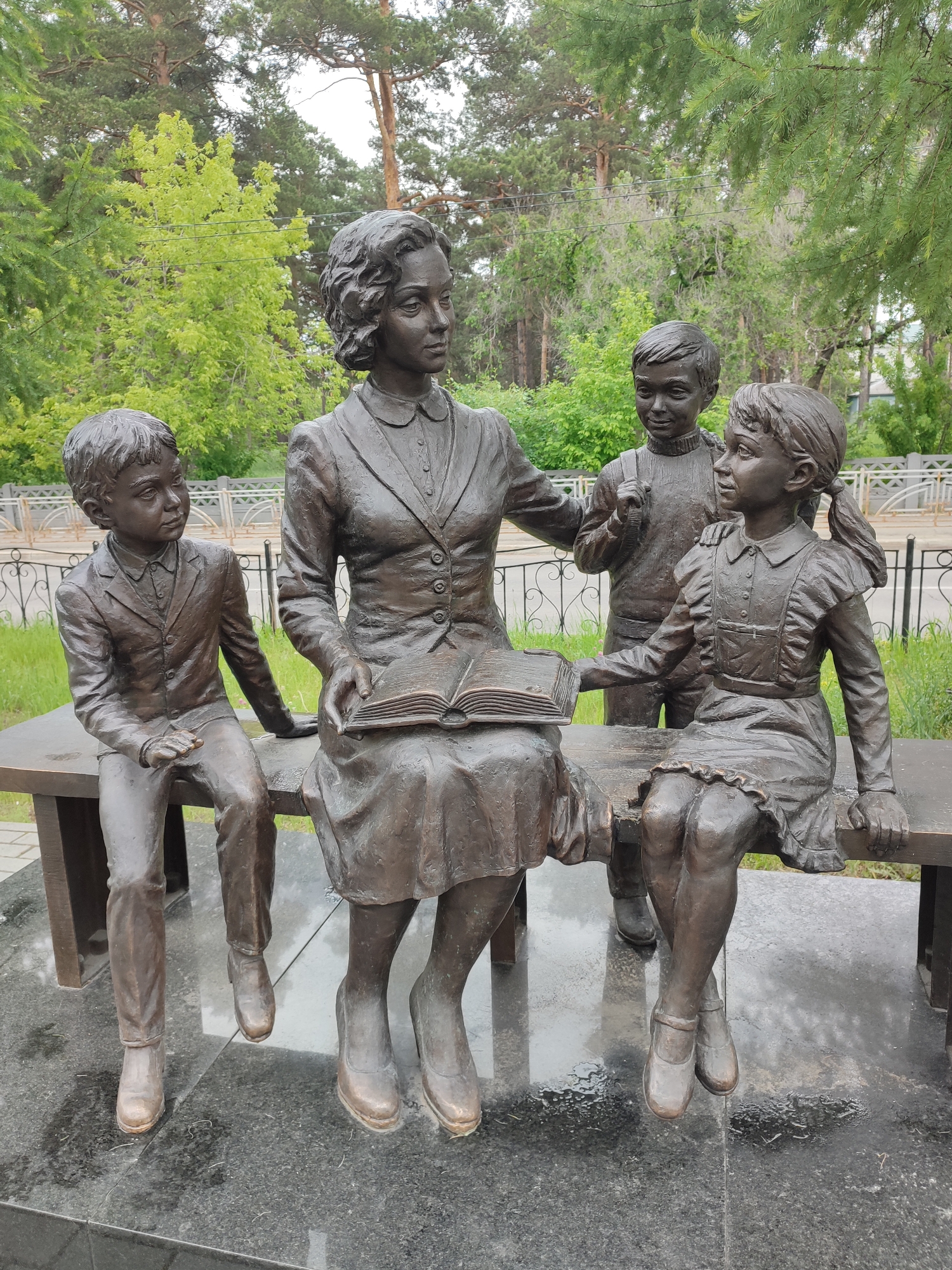 This bronze sculpture, located in the city of Angarsk, Russia, depicts a woman, presumably a teacher, reading a book to three children. The woman is seated on a bench, holding an open book in her lap. Two children, a boy and a girl, are seated beside her, also on the bench, facing her. A third child, a boy, stands behind the seated girl, with his hand resting on her shoulder. All figures are rendered in a realistic style, and the sculpture appears to be situated in an outdoor public space, possibly a park or square, with trees and a fence visible in the background. The ground beneath the sculpture is paved with dark, reflective stone, and there are wet patches, suggesting recent rain.