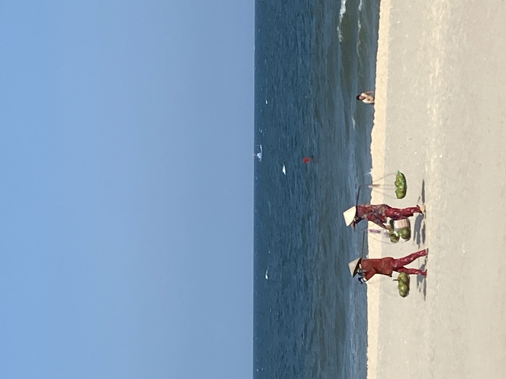 The image depicts a coastal scene at a beach in Phu Thuy Ward, Vietnam, under a clear, light blue sky. In the foreground, two individuals dressed in traditional Vietnamese attire, including conical straw hats (nón lá), walk from left to right across the light-colored sand. Each carries a bamboo pole balanced across their shoulders, from which two baskets are suspended, laden with numerous green, spherical fruits, likely coconuts. They cast distinct shadows on the sand.
The midground features the dark blue ocean with gentle waves breaking near the shore. To the right, a single individual is partially submerged in the shallow water, appearing to be sitting or wading, facing towards the beach. In the distance, a small white boat is visible on the horizon line, and closer in the water, a small red buoy or marker is present. The overall environment is open and uncrowded.