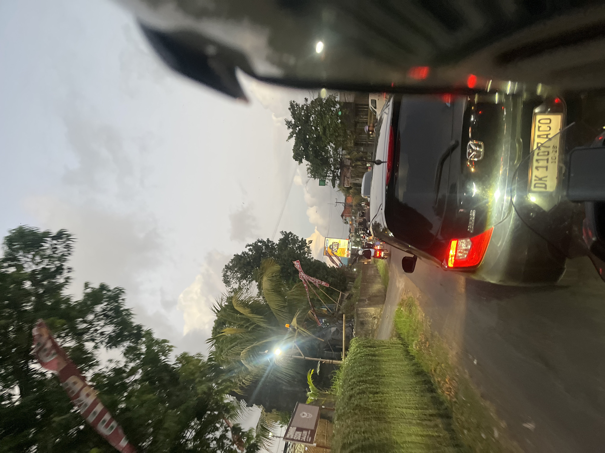 The image captures a street scene in Mengwi, Indonesia, likely during the evening. The perspective is from the viewpoint of someone on a motorcycle, evidenced by the helmet in the foreground and the handlebars. A dark-colored car, possibly a Mazda, is in the immediate foreground, with its license plate "DK 1107 ACO" visible. The car's brake lights are illuminated.
The road is lined with lush green vegetation on the left, possibly a hedge or hanging plants. Several vehicles, including motorcycles and cars, are present on the road, indicating traffic. One motorcycle in the middle distance has its headlight on. Further down the road, there are buildings and trees, with a billboard visible that says "FORTE". A street lamp with an orange light is also visible. The sky is cloudy, suggesting overcast weather or the approach of evening. Red and white flags or banners are strung between poles or trees in the background. The overall impression is of a typical street scene in a populated area of Mengwi, Indonesia, with moderate traffic and a tropical environment.