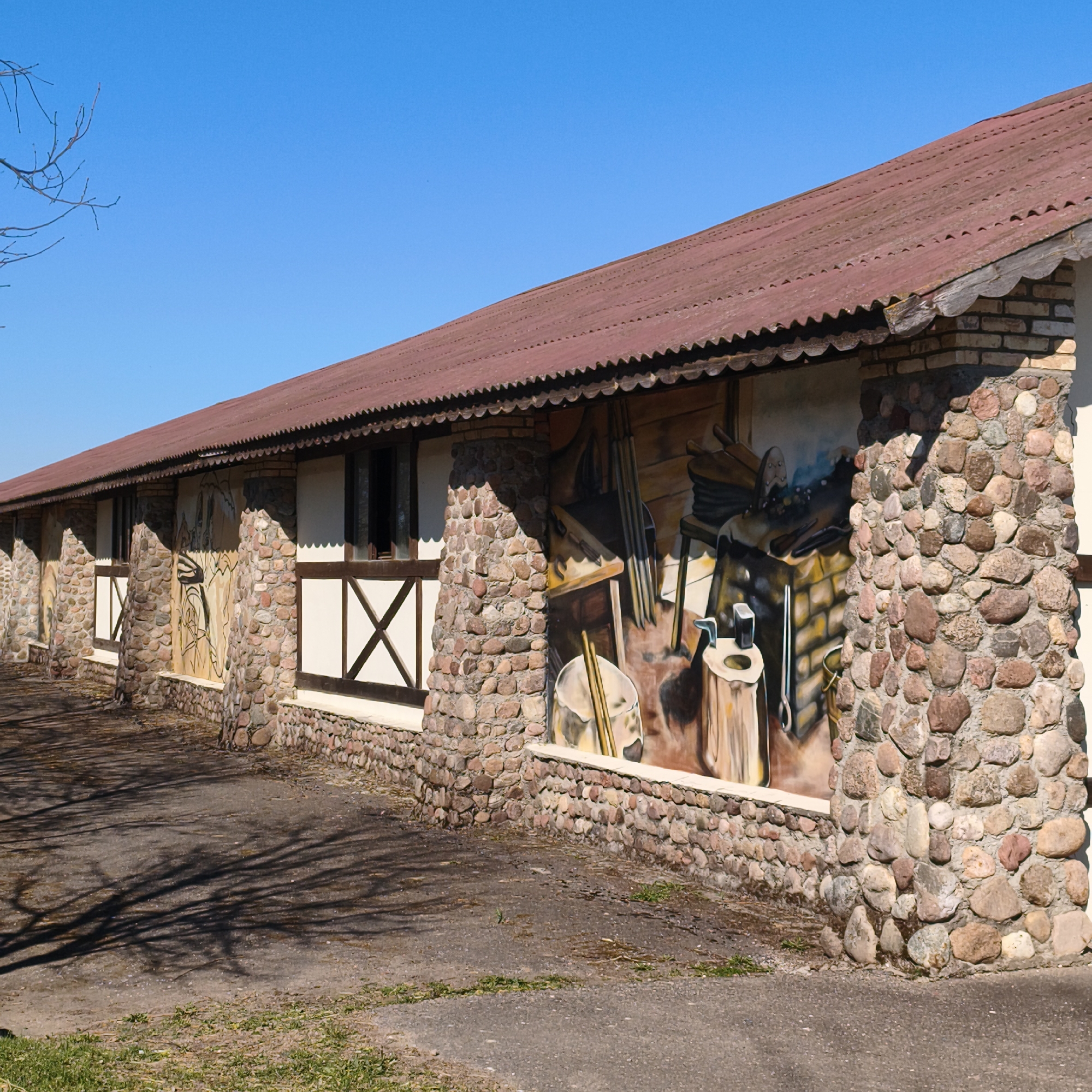 The image captures the side of a low, elongated building in Капцёўскі сельскі Савет, Belarus. The building has a distinctive architectural style, featuring alternating sections of whitewashed walls with dark brown half-timbered details and stone pillars constructed from rounded river rocks. Between the sections with the window and half-timbered construction are sections with paintings of blacksmith tools and workbenches. The building is topped with a rusty brown corrugated roof. The foreground is a paved area with scattered grass and the shadows of bare tree branches cast upon it. The sky above is clear and bright blue.