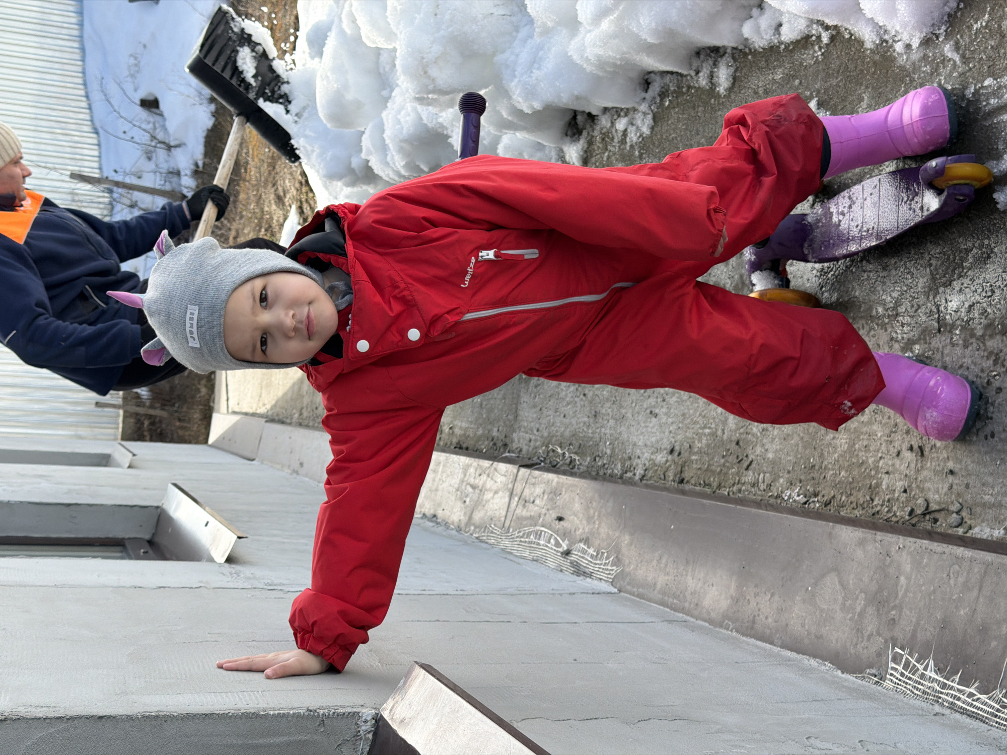The image depicts an outdoor scene in the city of Chaikovsky, Russia, featuring two individuals amidst snow. In the foreground, a young child, estimated to be between three and five years old, stands facing the camera. The child is dressed in a red full-body snowsuit, a grey knitted hat with pink unicorn-like ears, and pink rubber boots. The child's left hand is pressed flat against a light grey, textured stucco wall. A purple kick scooter with yellow wheels is partially visible beneath the child's legs, resting on a concrete surface. This surface is partially covered with white snow and meltwater. In the background, positioned to the right of the child, an adult is visible, wearing a dark blue jacket with an orange high-visibility stripe on the shoulder, a light-colored beanie, and dark gloves. This adult is actively engaged in shoveling white snow with a black shovel that has a wooden handle. The background also shows a corrugated metal fence or wall and patches of bare ground. The stucco wall features two visible metal window sills.