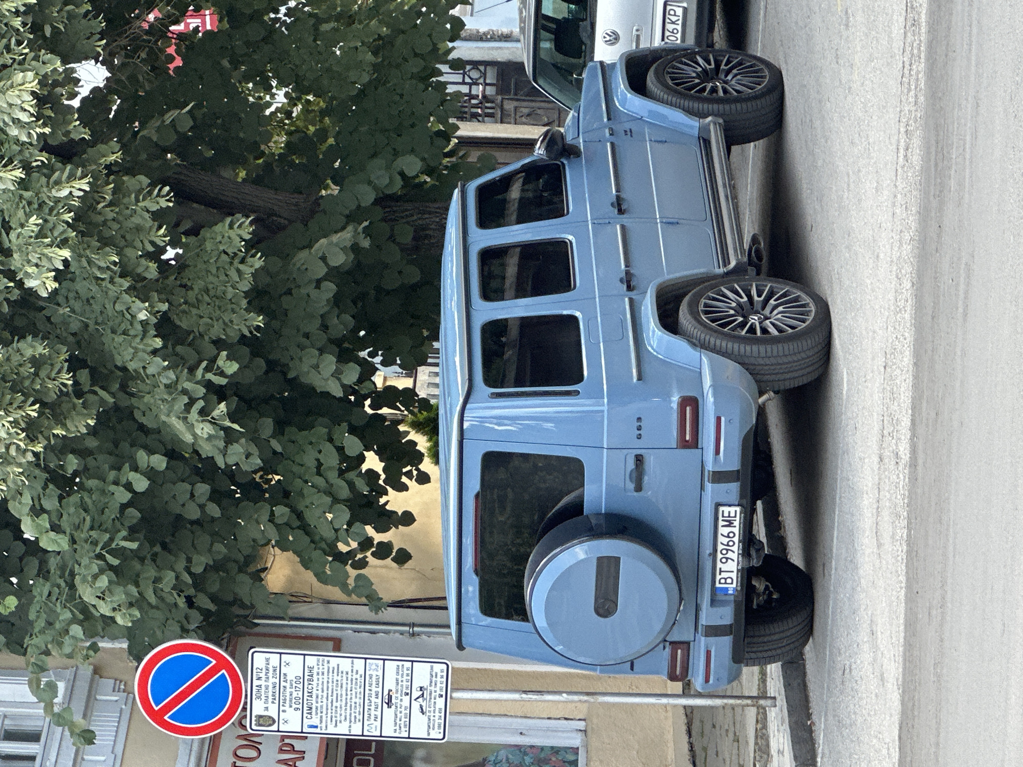 The image shows a light blue Mercedes-Benz G-Class G63 parked on a city street in Veliko Tarnovo, Bulgaria. The vehicle is facing to the right, with its rear end prominently featured. A spare tire is mounted on the rear door. A Bulgarian license plate reading "BT 9966 ME" is visible.
To the left of the vehicle, a parking sign with a red circle and a diagonal bar across a blue background indicates a "No Stopping" zone. Below this, a detailed sign with Bulgarian text provides information about parking regulations, including operating hours and payment methods. Text on the sign mentions "PAY FAST AND EASILY" and a phone number for inquiries.
In the background, a large tree with dense green foliage provides shade. To the right of the G-Class, a portion of a silver car is visible, with a license plate that appears to read "06 KP". The street is paved with asphalt, and a sidewalk is present. The architecture in the background suggests an urban setting. No people are visible in the image. The scene is captured during daylight hours, with ample natural light.