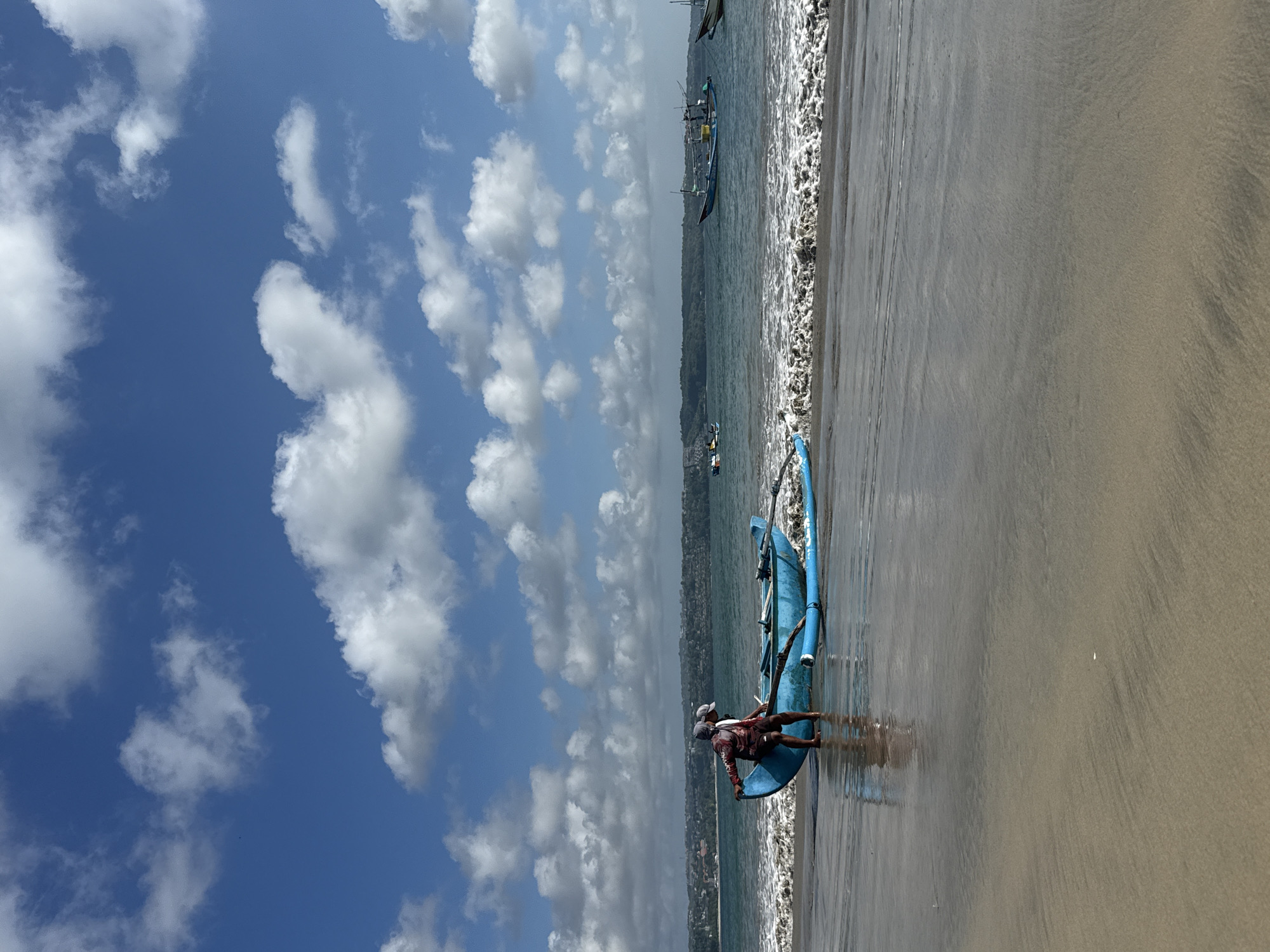 A medium shot, captured vertically, showcases a beach scene in Kuta, Indonesia, under a partly cloudy sky. The sky, filled with puffy white clouds against a backdrop of blue, dominates the upper half of the frame. The lower half is composed of wet sand meeting the ocean. A blue boat rests on the shore, with two individuals positioned near it. One is bent over the boat. The second individual stands behind, facing the horizon. In the distance, additional boats are visible on the water near the shoreline.