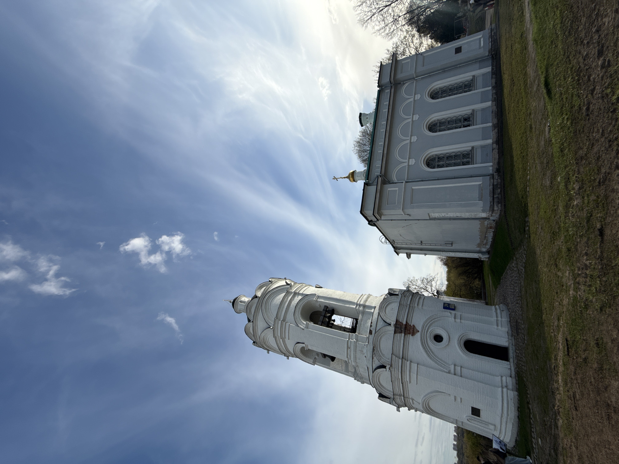 The image shows two buildings in Moscow, Russia, viewed from a low angle. A white bell tower stands prominently in the foreground, against a backdrop of blue sky with streaky clouds. The tower features arched openings and a series of tiered sections. Behind the tower and slightly to the right is a light gray building with arched windows and a golden dome topped with a cross. The ground around the buildings is covered in green grass. The photo is rotated counter-clockwise.