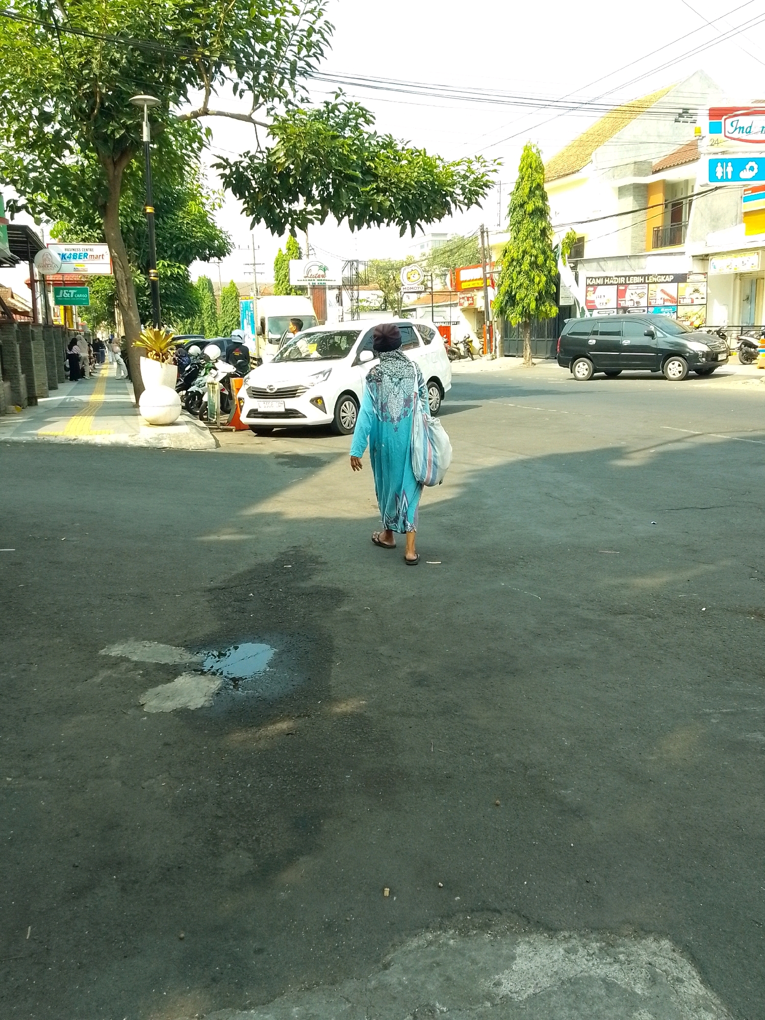 A medium shot shows a person walking away from the camera on a street in Jember, Indonesia. The person is wearing a long, blue, patterned garment and carrying a bag. They are walking on asphalt with visible tire marks and a puddle of water on the left side of the frame. In the background, there is a white car and a black SUV parked on the street. Buildings with signs are visible on the right side of the street, including a sign for "Indomaret" and another for "KAMI HADIR LEBIH LENGKAP." Trees with green foliage are also present, one of which is large and near the left side of the image, and another tall, slender green tree is in the middle ground. The sky is bright and appears to be daytime. A sidewalk with yellow tactile paving is on the left side of the street, with several parked motorcycles and a few people walking on it.