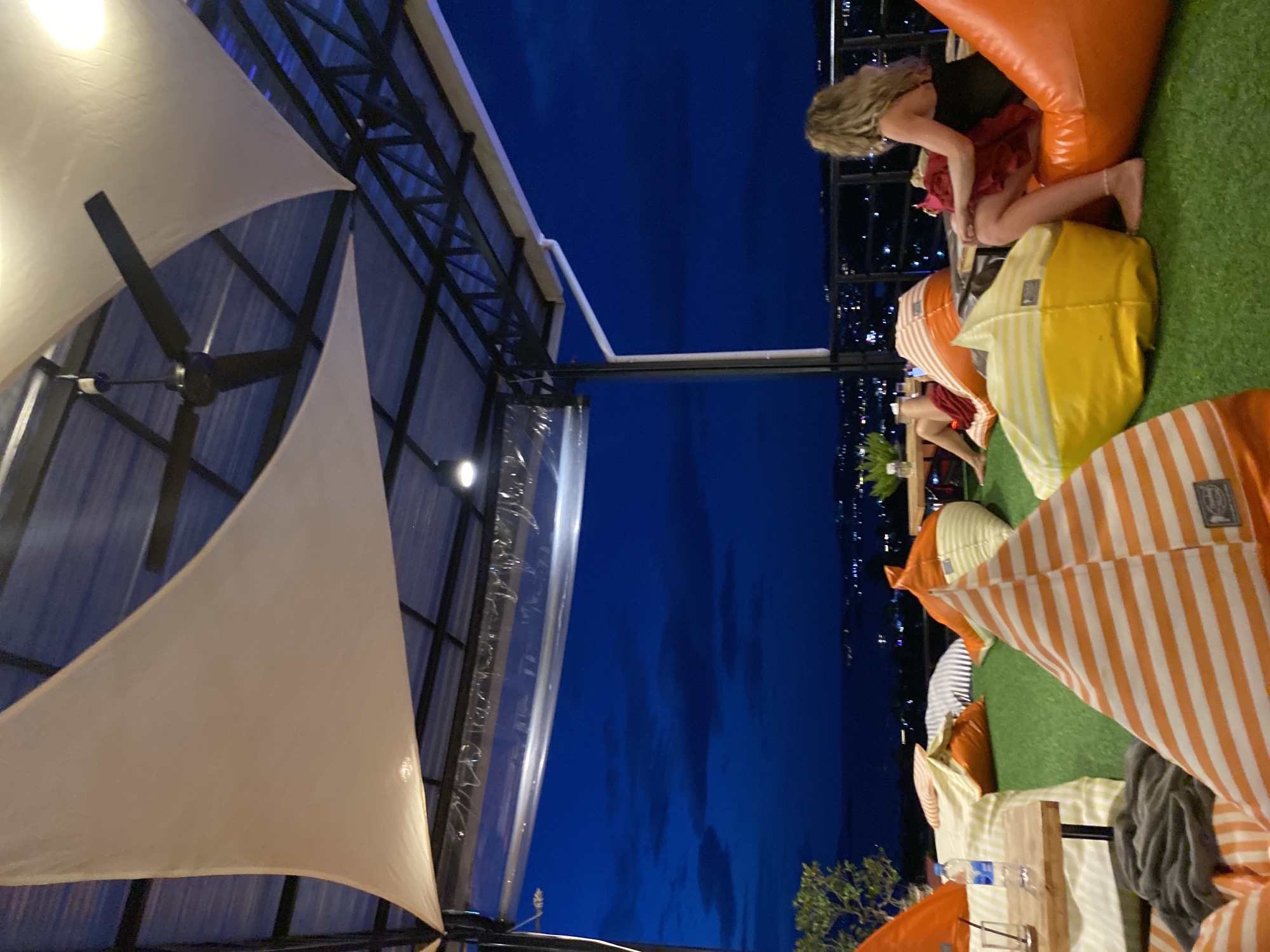 This image shows a rooftop outdoor lounge area in บ้านบางรักษ์, Thailand. The scene is shot at night, with a view of the city lights in the background. The seating consists of colorful beanbag chairs arranged on artificial grass. A blonde-haired woman is sitting in an orange beanbag chair. Other individuals are visible in the background. The area is partially covered by white sail-like awnings supported by a black metal frame. A ceiling fan is attached to the frame. There is also clear plastic sheeting that can be lowered to block the wind. A table with water bottles and glasses can be seen among the beanbag chairs.