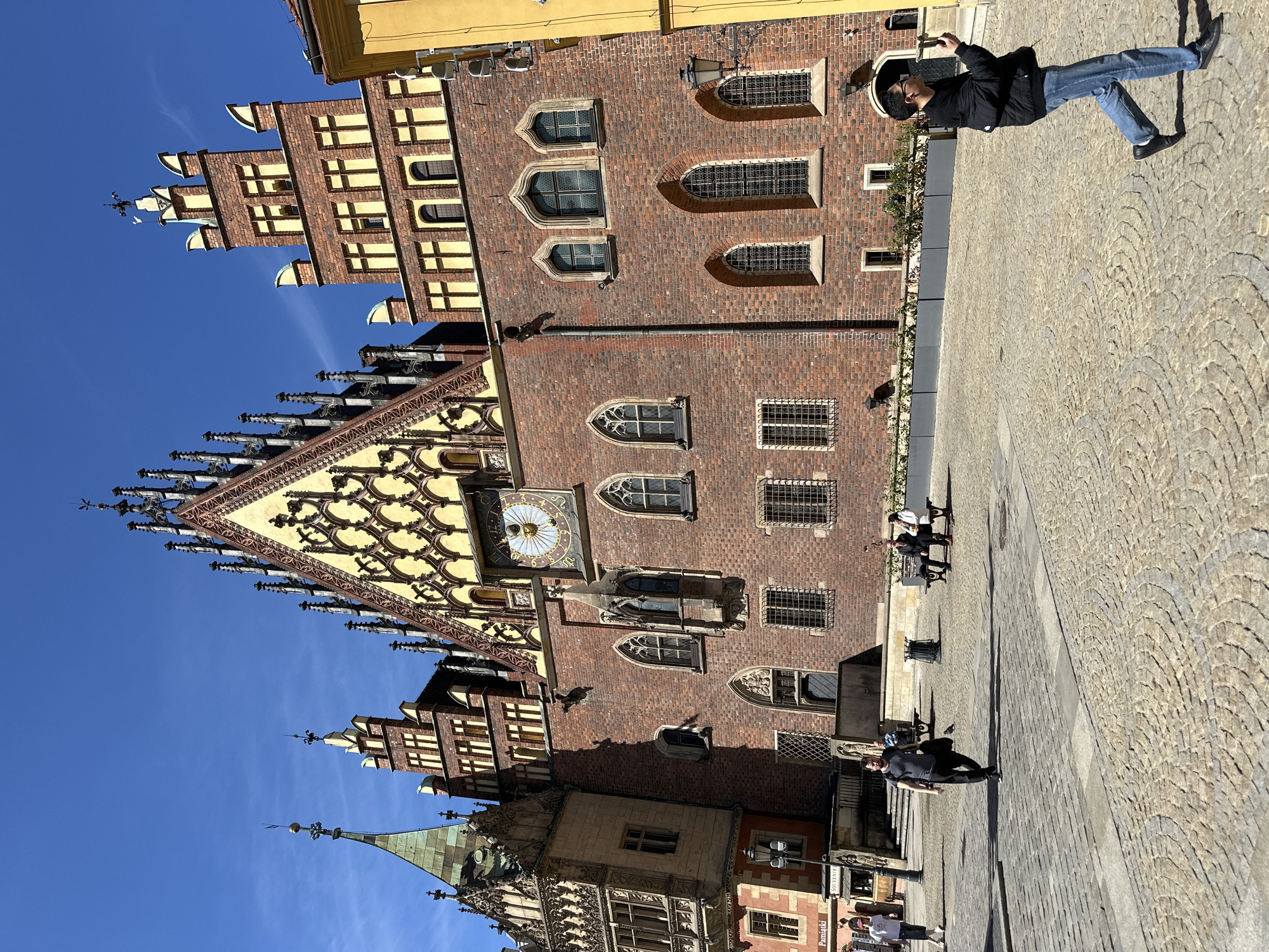 This image shows the historical architecture of Wrocław, Poland. The prominent building is the Old Town Hall, known for its Gothic style. The photo captures the detailed façade, including the clock, windows, and ornate decorations. The sky is clear and blue. Several people are walking around the paved square. In the foreground, a person is wearing a black jacket and blue jeans.