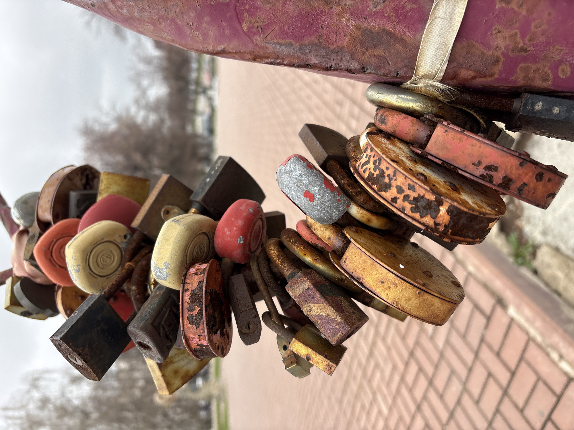 The image depicts a close-up view of a cluster of love locks attached to a structure. The locks vary in size, shape, and color, with many exhibiting signs of rust and wear. There are round, square, and rectangular locks in shades of red, gold, silver, black, and cream. They are interlocked, creating a dense mass of metal. A worn, rust-colored beam supports the locks, and a white ribbon or strap is tied around it. The blurred background suggests an outdoor environment, possibly a bridge or railing in Ulyanovsk, Russia. A brick surface and bare trees can be vaguely discerned.