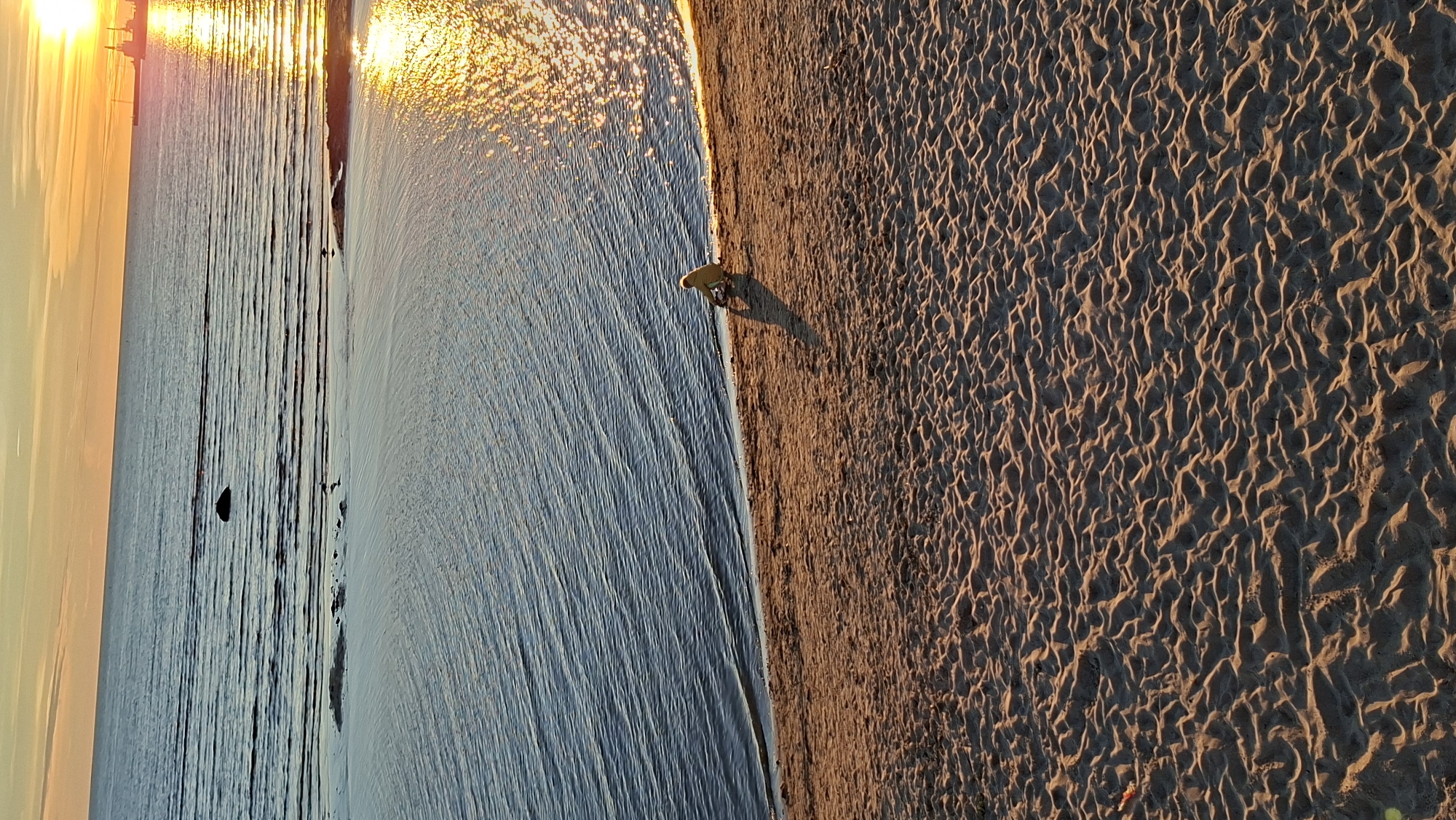 A person wearing a yellow jacket walks along the edge of the water on a sandy beach at sunset. The sun is low in the sky, casting a warm golden glow on the scene and reflecting brightly on the rippling water. The sand is textured with small waves and ripples, indicating it has been recently disturbed by the tide or wind. In the distance, across the water, a faint silhouette of structures can be seen, possibly part of a coastline or a city skyline. The overall atmosphere is serene and peaceful, suggesting a tranquil evening by the sea in Tallinn, Estonia. There is no visible text in the image.