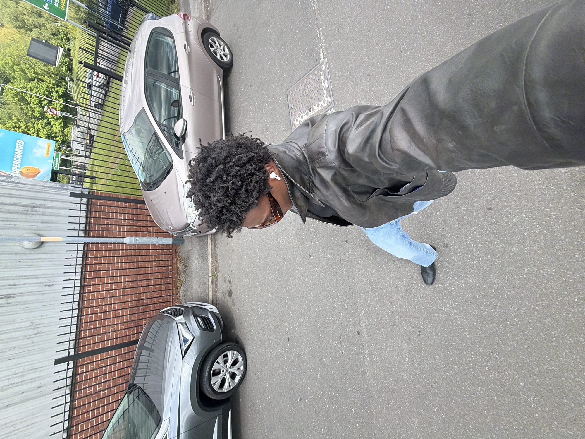 The image shows a person standing on an asphalt road next to a brick wall and a metal fence. Two cars are parked alongside the road: a silver Peugeot and a grey SUV. The person is wearing a black leather jacket, light blue jeans, and black shoes. The person has afro-style hair and is wearing sunglasses and an earbud in their right ear. The person is holding the camera in their hand, taking a selfie. A sign in the background has text. The image location could possibly be Manchester based on context clues.