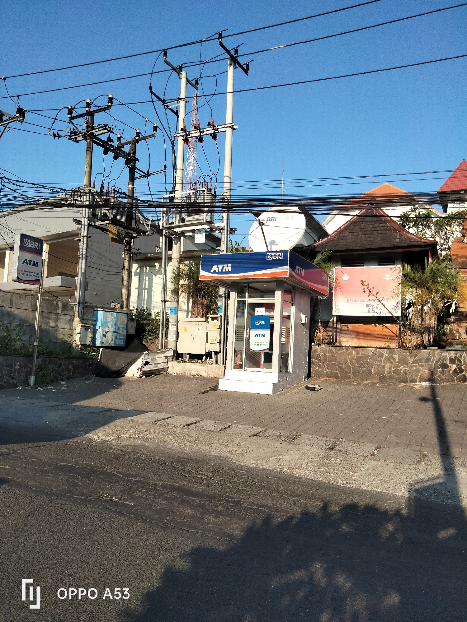 The image shows a street scene in Denpasar, Indonesia, under a clear blue sky. A BRI ATM booth is prominently featured on the right side of the frame. The ATM booth is modern, with a blue and orange sign indicating "BRI ATM." To the left of the ATM booth, another BRI ATM sign on a pole is visible. Electrical poles with numerous wires and a transformer are situated behind the ATM booth, indicating urban infrastructure.
In the background, several buildings are visible, including one with a traditional Balinese-tiled roof. Some vegetation, including palm trees, adds to the tropical environment. The ground is paved with interlocking bricks and asphalt, with some debris and shadows cast by the sun, suggesting it is daytime. A large satellite dish is mounted on top of one of the buildings. A billboard with text and an image is mounted on a stone wall. The scene appears to be a typical street view in a commercial or residential area of Denpasar.