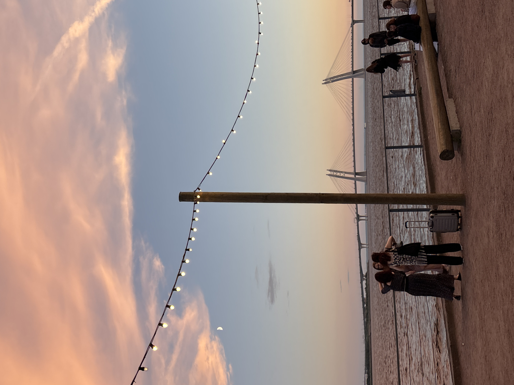 The image depicts a waterside promenade in Saint Petersburg, Russia, during the golden hour. The sky is painted with hues of pink and orange from the sunset, contrasting with the clear blue sky above. A string of lights is draped across the scene, casting a warm glow. In the distance, the impressive cable-stayed bridges of Saint Petersburg are visible over the Gulf of Finland.
On the promenade, several individuals are present. Three women are clustered together near the water's edge, with one holding a suitcase. Further down the promenade, a group of people are seated on a long wooden bench, and others are standing and conversing. The ground is covered in a reddish-brown gravel. The water is calm, reflecting the colors of the sky. A crescent moon is faintly visible in the upper left portion of the sky.