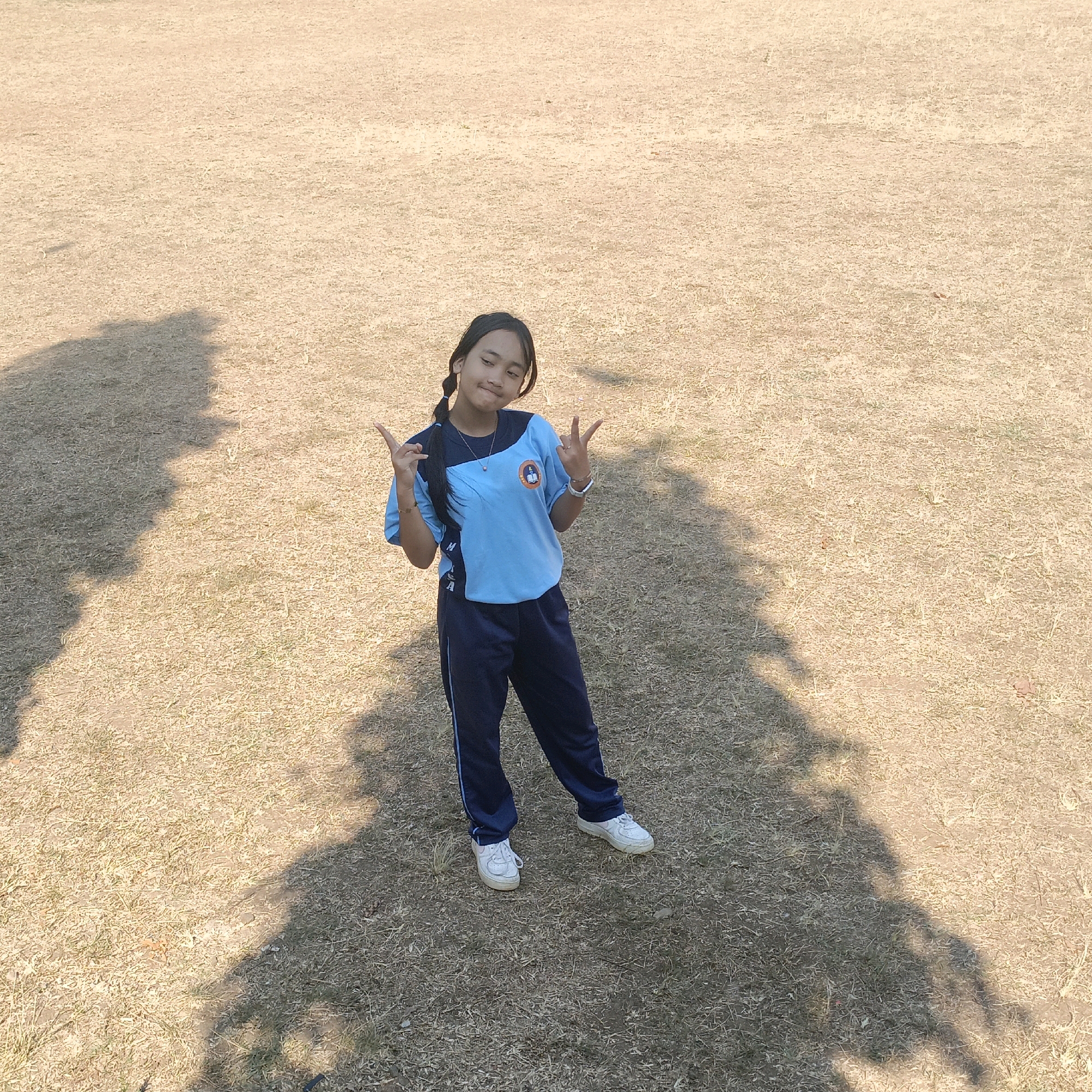 A young girl with braided hair is standing in a dry grassy field, possibly a park or an open area in Salatiga, Indonesia. She is wearing a light blue t-shirt with dark blue sleeves and a dark blue tracksuit bottom with a light blue stripe. On her t-shirt, there is a circular emblem with a figure and some text. She is also wearing white sneakers and a silver necklace. The girl is looking towards the camera with a slight smile and is making a "peace" sign with both hands. Long shadows from trees or structures are cast across the field. The ground is covered with short, dry, light brown grass.