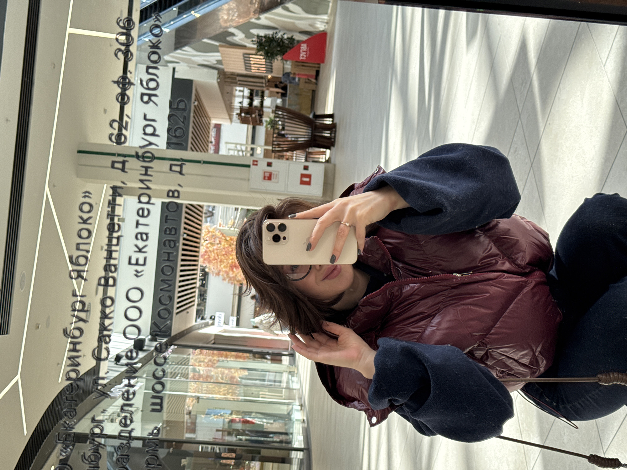 The image is a mirror selfie taken by a person in an indoor, brightly lit setting, likely a modern shopping mall or commercial building in Perm, Russia. The photograph is oriented 90 degrees clockwise from its natural upright position.
The main subject is a person, likely female, whose face is partially obscured by the light-colored smartphone (appearing to be an iPhone 14 Pro or 15 Pro) she holds to take the photo. She wears glasses, a burgundy puffer jacket, and dark blue fleece sleeves. A ring adorns a finger on the hand holding the phone, and dark nail polish is visible. Her short, dark hair frames her face, and a slight smile is discernible beneath the phone.
The background, visible through the large reflective surface (mirror or glass wall), showcases a modern interior with light-colored walls and flooring, suggesting ample natural or artificial light. Reflections show contemporary architectural elements, including wooden structures, what appear to be seating areas, and some potted plants.
Prominently reflected on the surface is reversed Cyrillic text, which provides location details. When read correctly, it displays: "«Екатеринбург Яблоко»" (Yekaterinburg Yabloko), an address "Сакко Ванцетти, д. 62-оф.-306" (Sacco Vanzetti, bld. 62, office 306), "разделение ООО «Екатеринбург Яблоко»" (division OOO "Yekaterinburg Yabloko"), and crucially, "Пермь шоссе Космонавтов, д. 1625" (Perm, Kosmonavtov highway, bld. 1625), confirming the given location context. Other faint text includes "VILAGE" (reflected upside down from a red sign) and "TONIBA".