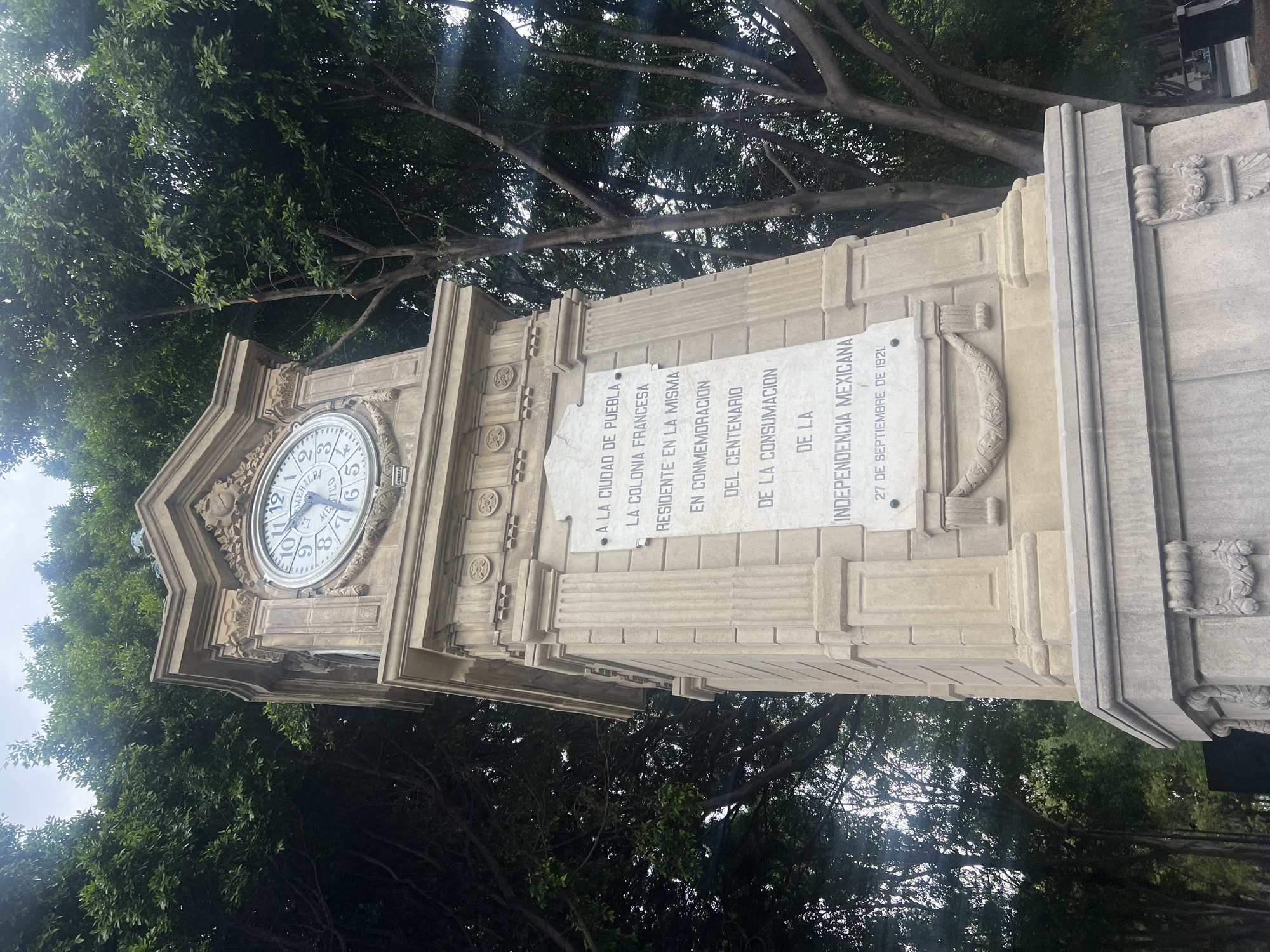 The image shows a stone clock tower with a clock face displaying the time as approximately 11:12. The clock face also bears the inscription "MERALBA MEXICO". Next to the clock is a plaque with Spanish inscriptions. The inscription translates to: "TO THE CITY OF PUEBLA. THE FRENCH COLONY RESIDENT IN THE SAME IN COMMEMORATION OF THE CENTENARY OF THE CONSUMMATION OF THE MEXICAN INDEPENDENCE 27TH OF SEPTEMBER OF 1921." The tower is surrounded by trees, and the sky is visible through the foliage.