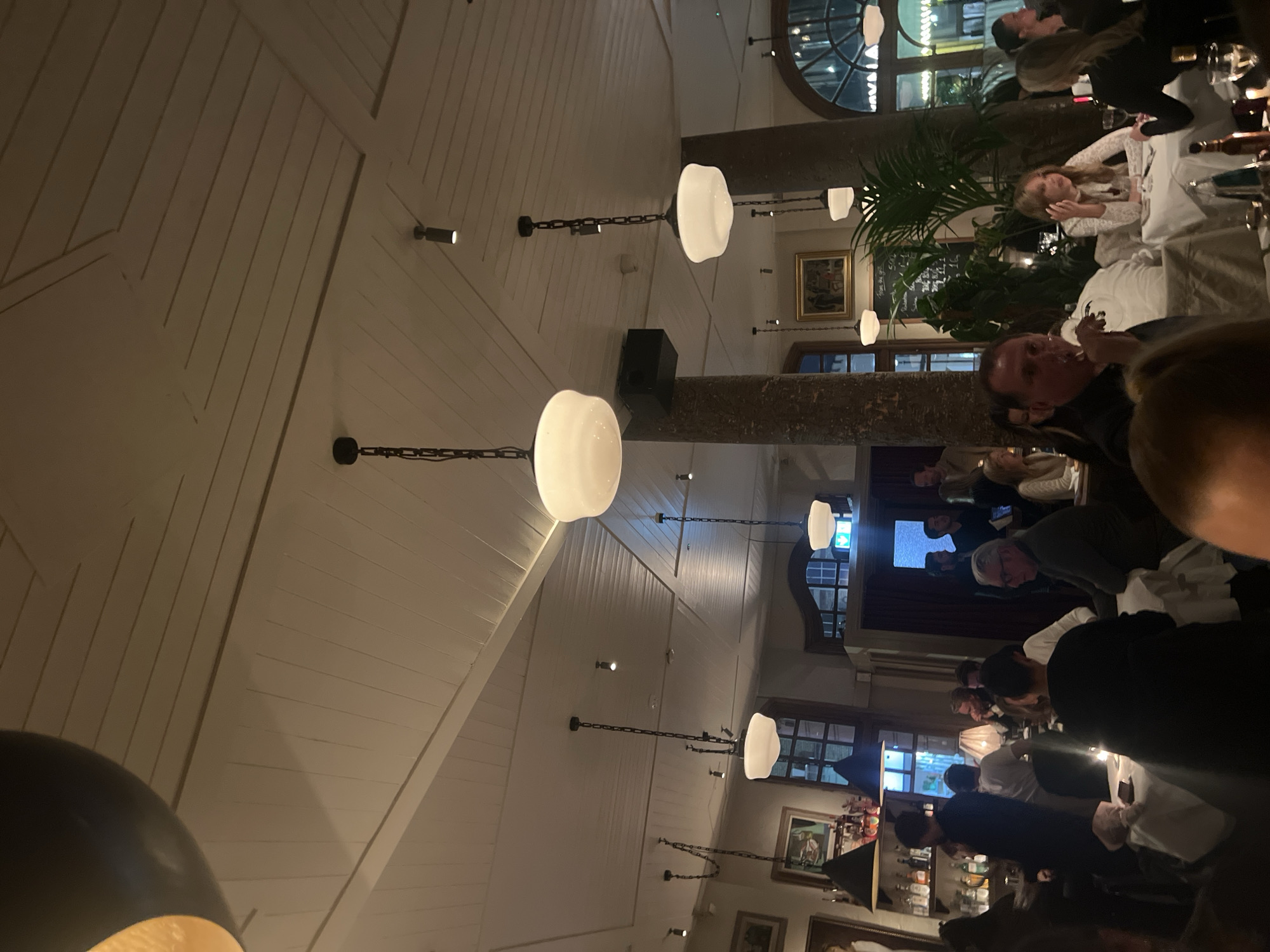 The image depicts the interior of a restaurant, likely in London, United Kingdom, based on the architectural style and ambiance. The restaurant features a high, gabled ceiling with white wooden paneling, punctuated by a series of chain-hung pendant lights with globe shades. Several diners are seated at tables, engaged in conversation and dining. The tables are covered with white tablecloths and set with glassware. In the background, there are windows and framed artwork on the walls. Architectural columns are visible, potentially supporting the structure. The lighting is warm and ambient, creating a cozy atmosphere.